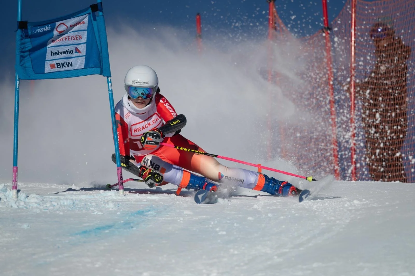A skier crashes while rounding a gate during an alpine ski race in snowy conditions, with snow spray and safety netting in the background.