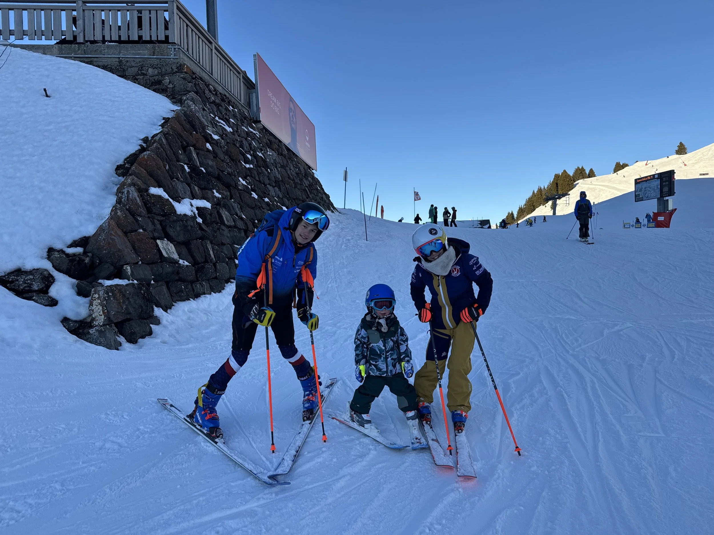 A group of children and adults in ski gear standing on a snowy slope at a ski resort, with a snow-covered mountain and ski lift in the background.