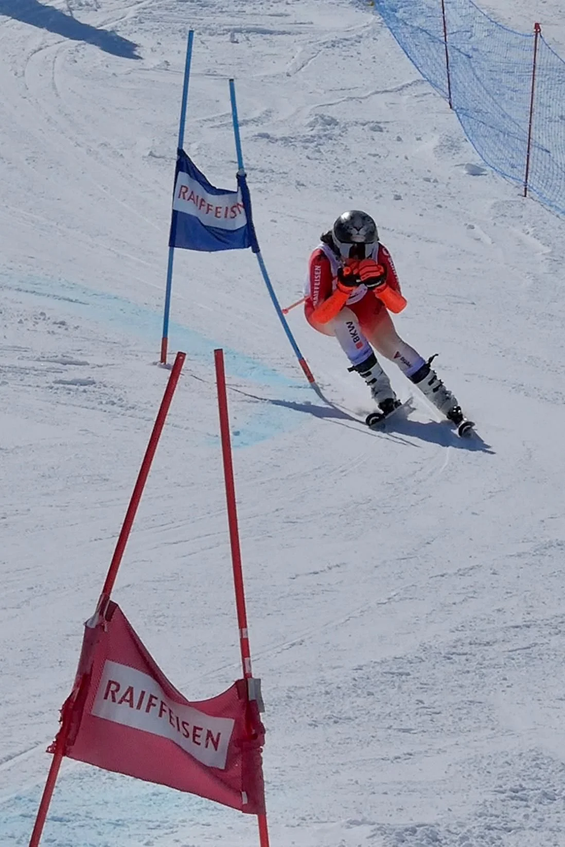 A skier wearing an orange and white suit, helmet, and goggles is racing on a snowy slope, navigating around red and blue gate poles with the word 'Raiffeisen' on the flags.