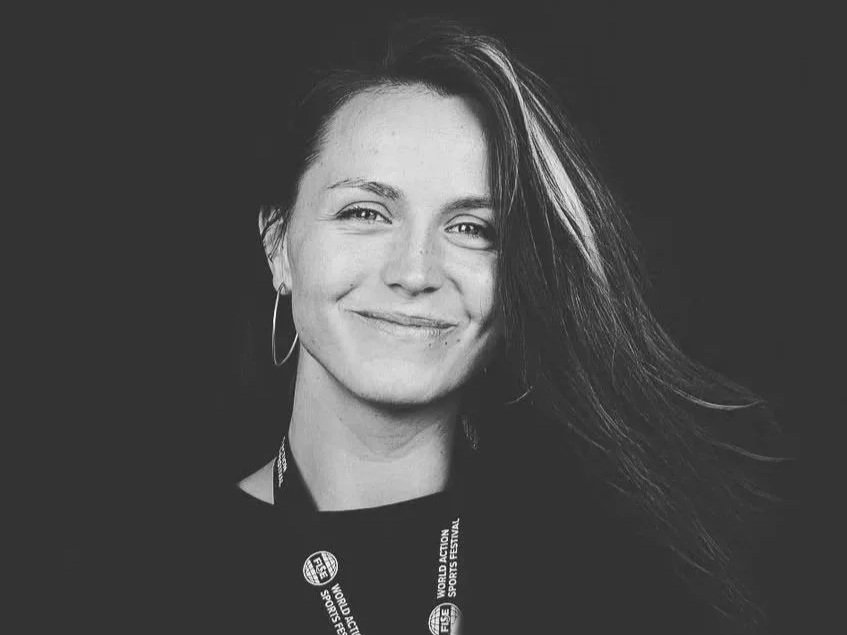 Black and white portrait of a woman smiling, with long hair flowing to the right, wearing hoop earrings and a lanyard that reads 'World Action Sports Festival'.