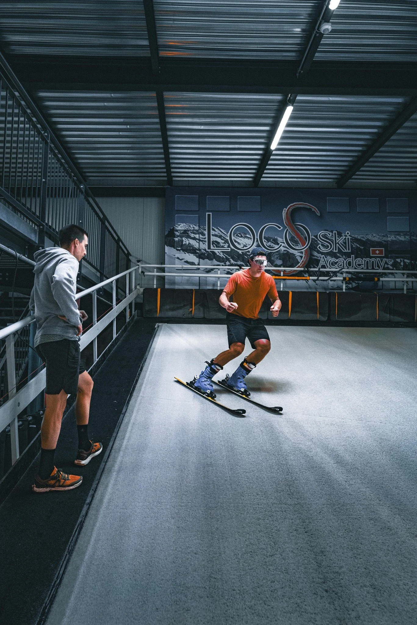 An indoor ski training facility featuring a skier in orange athletic wear maneuvering on synthetic snow. A person in a hoodie observes from the side, and a sign on the wall reads 'Loco Ski Academy' with mountain imagery and Swiss flag.