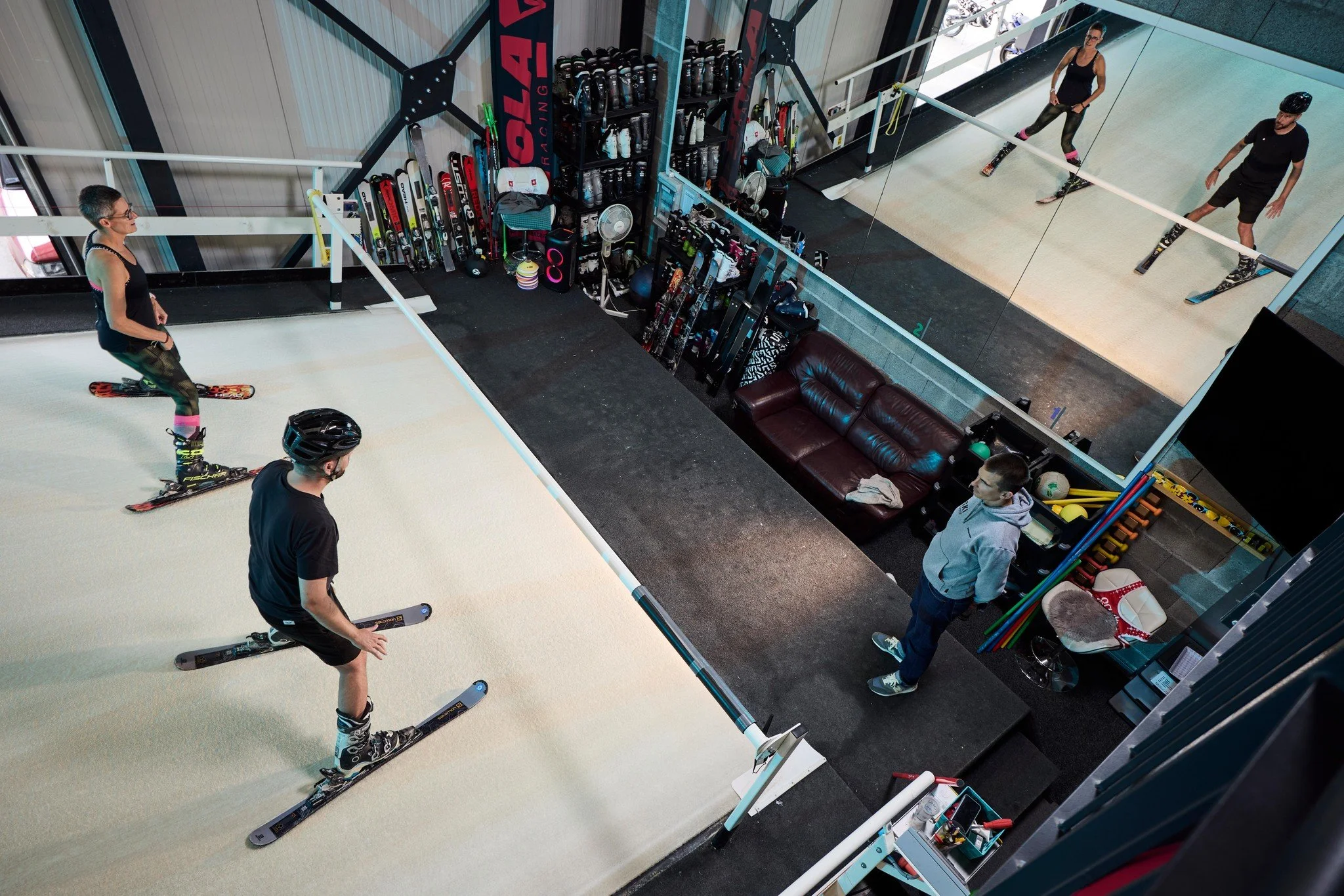 People preparing for skiing in an indoor ski training area, with skis and gear stored on racks and a large mirror.