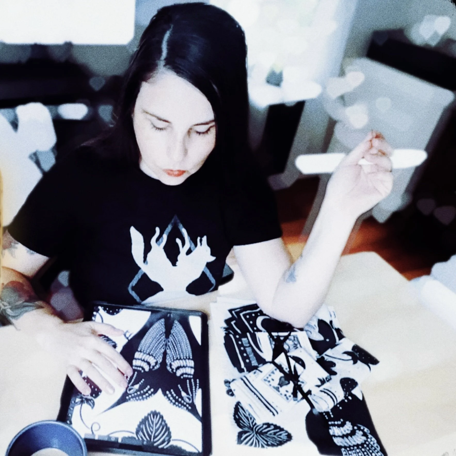 A woman with black hair, pale skin, and tattoos on her arms, sitting at a table with her artwork and products.  She is wearing a black t-shirt with a coyote design. Her eyes cast downward, and she is holding an apple pen in one hand.