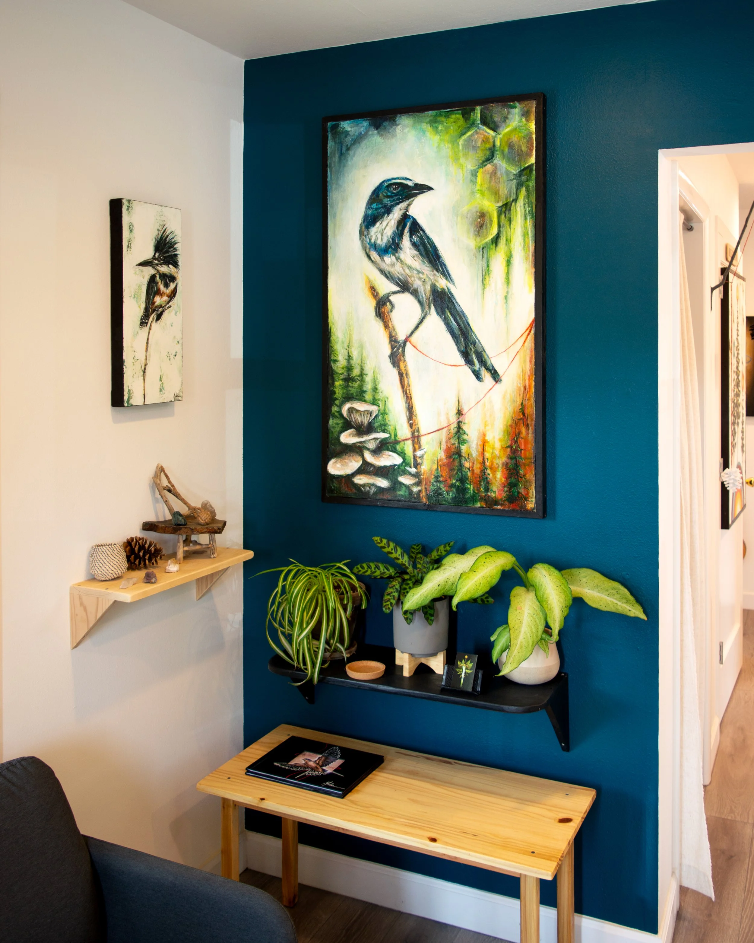 Interior of a room featuring a blue accent wall with a large bird painting, smaller bird artwork, and a shelf with plants, decorations, and a table with magazines.