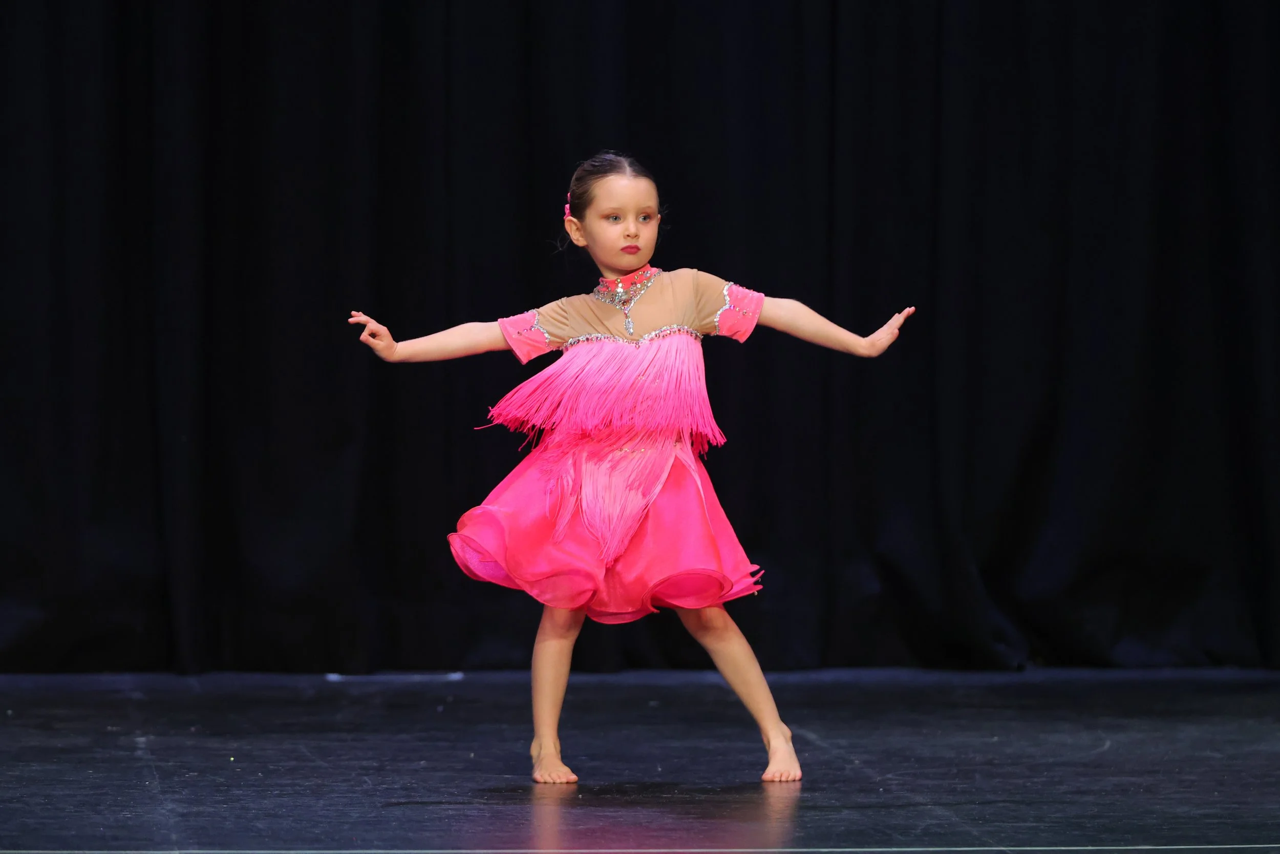 Young girl performing a dance on stage wearing a bright pink dress with fringe details and rhinestone embellishments, with a black curtain backdrop.