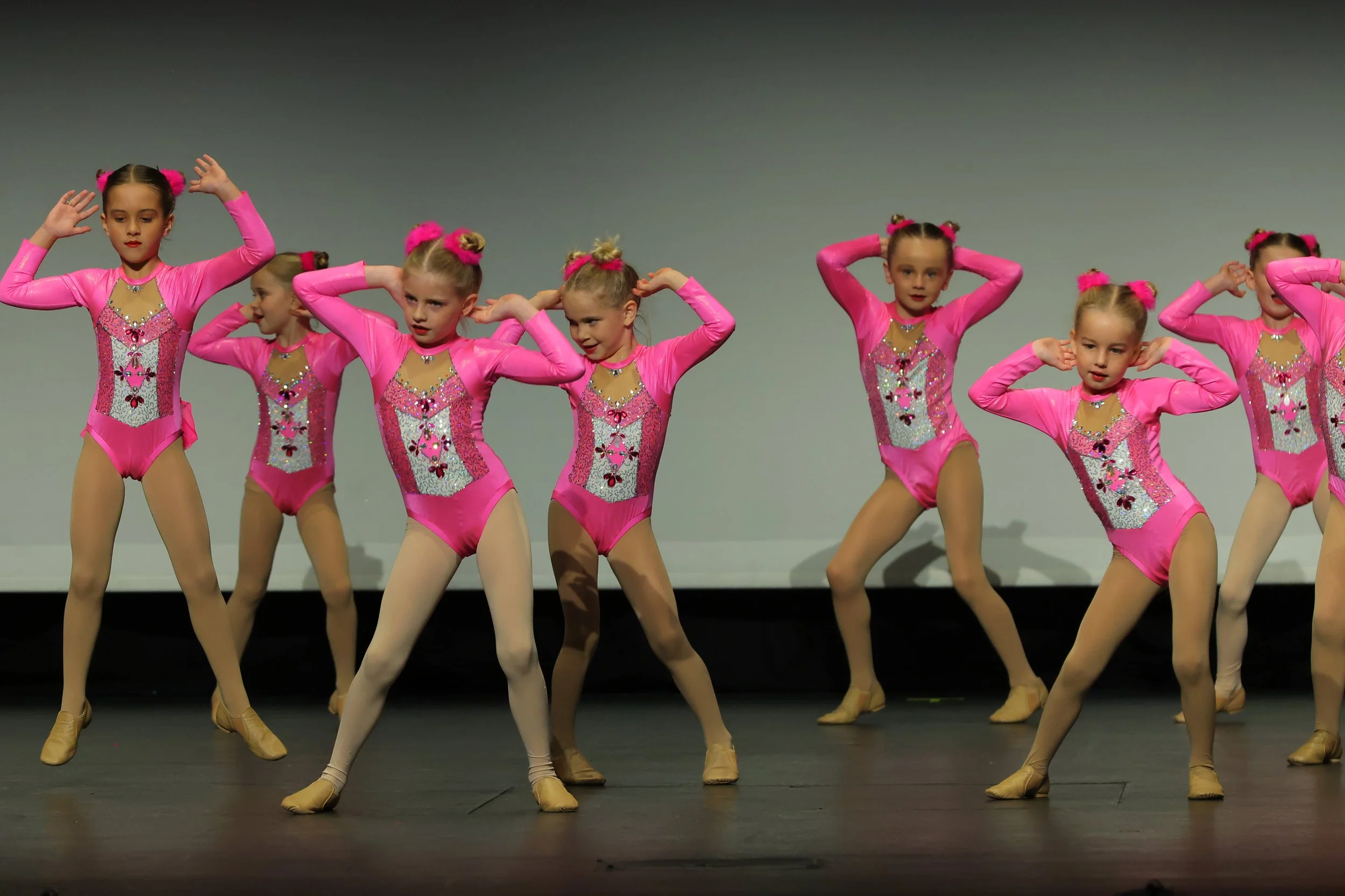 Group of young girls performing a ballet dance on stage, wearing pink costumes with sparkly details and beige ballet shoes.