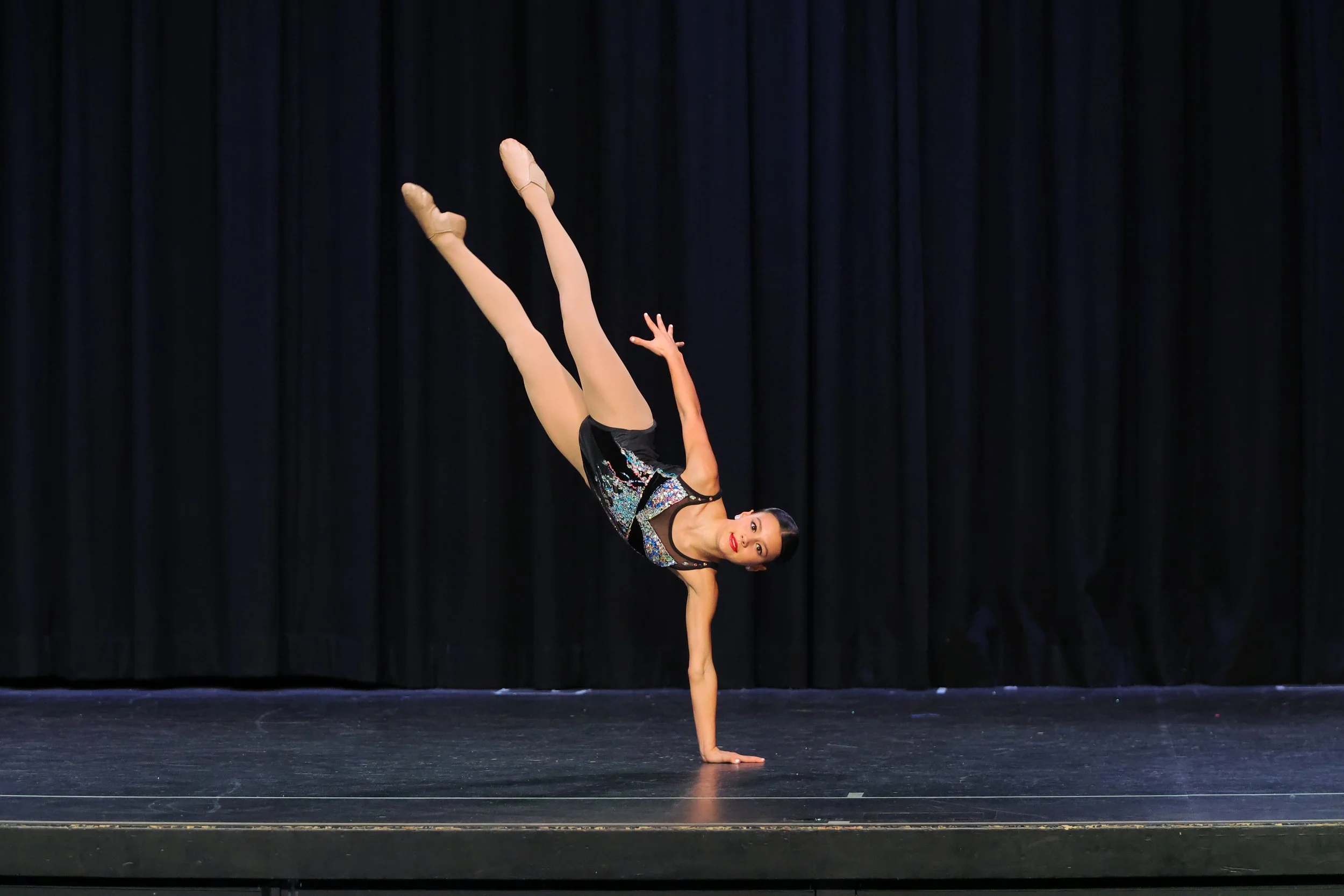 A female gymnast performs a handstand on stage with a black curtain backdrop, wearing a sparkly leotard.