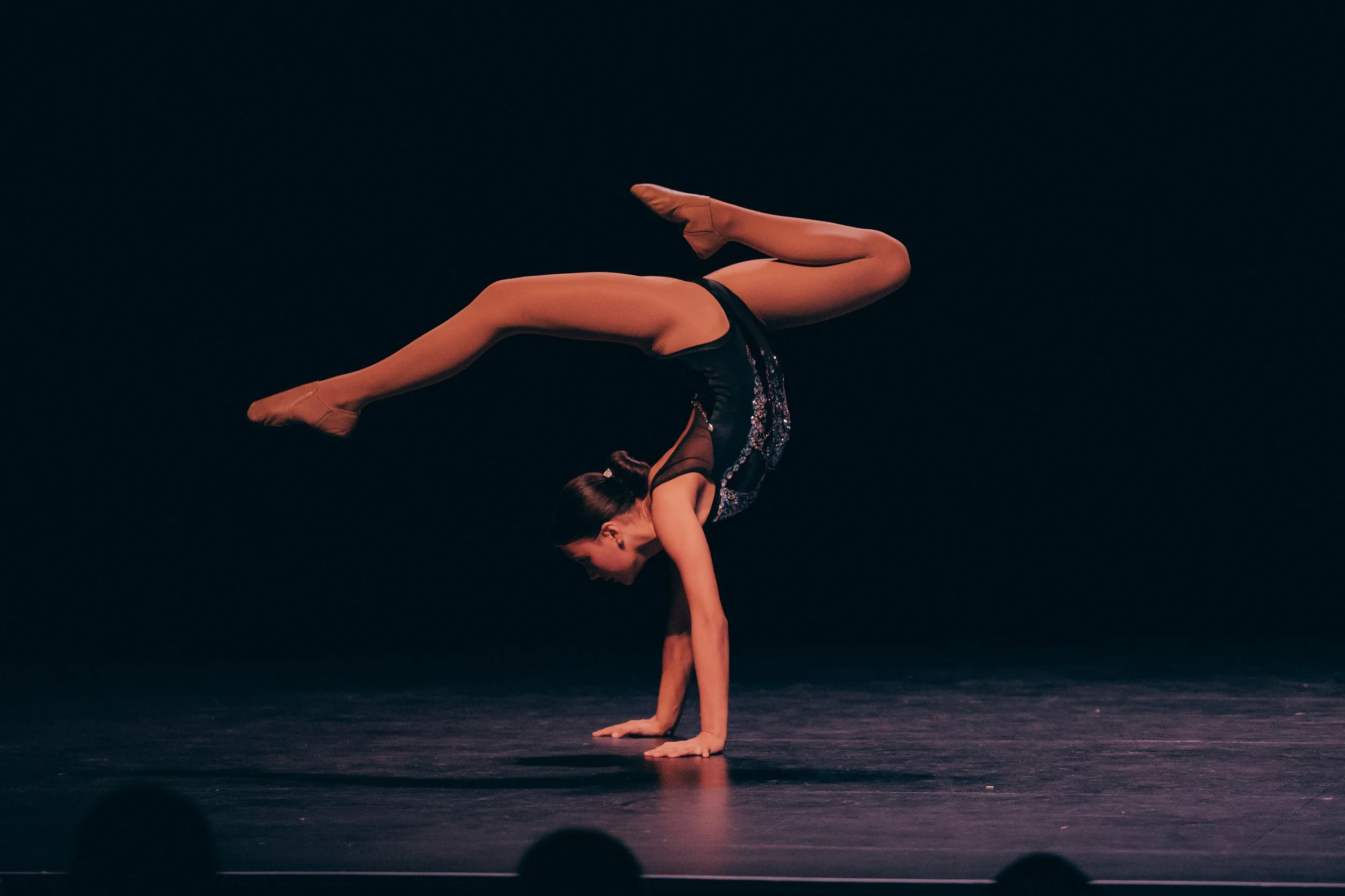 A female performer balancing on her hands in a handstand on a dark stage, wearing a black costume with shiny embellishments, with a black background and some audience silhouettes at the bottom.