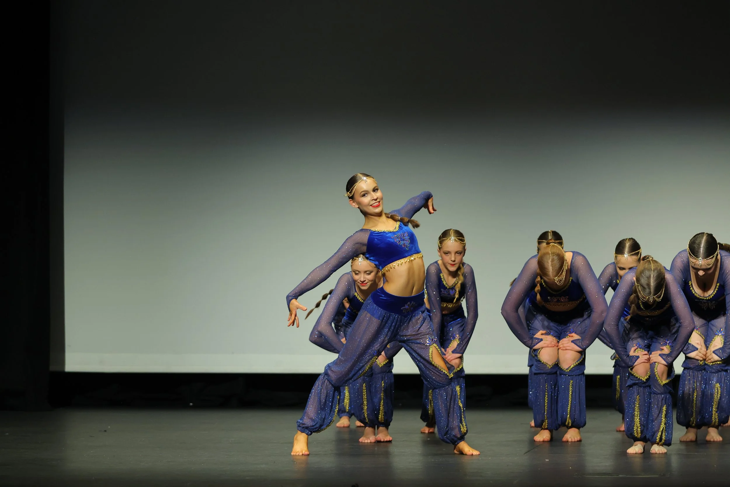 A group of young female dancers in blue costumes performing on stage, with one girl standing confidently in front while others bend forward.