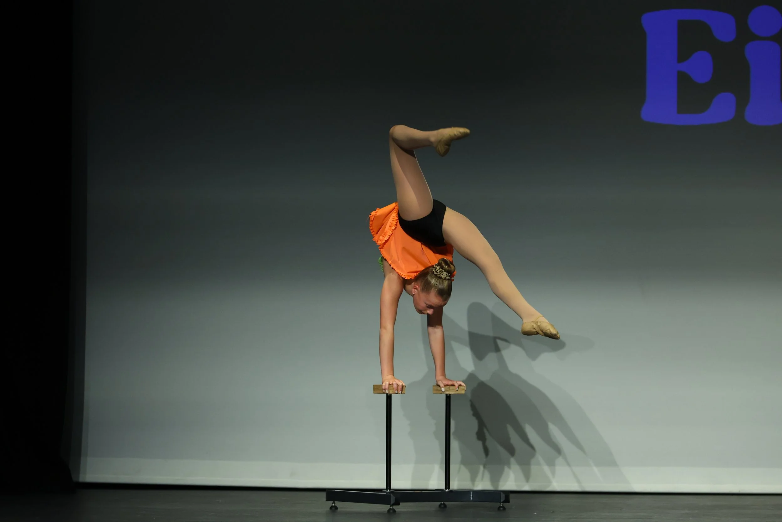 A female acrobat performing a handstand on parallel bars on stage, wearing an orange and black costume.
