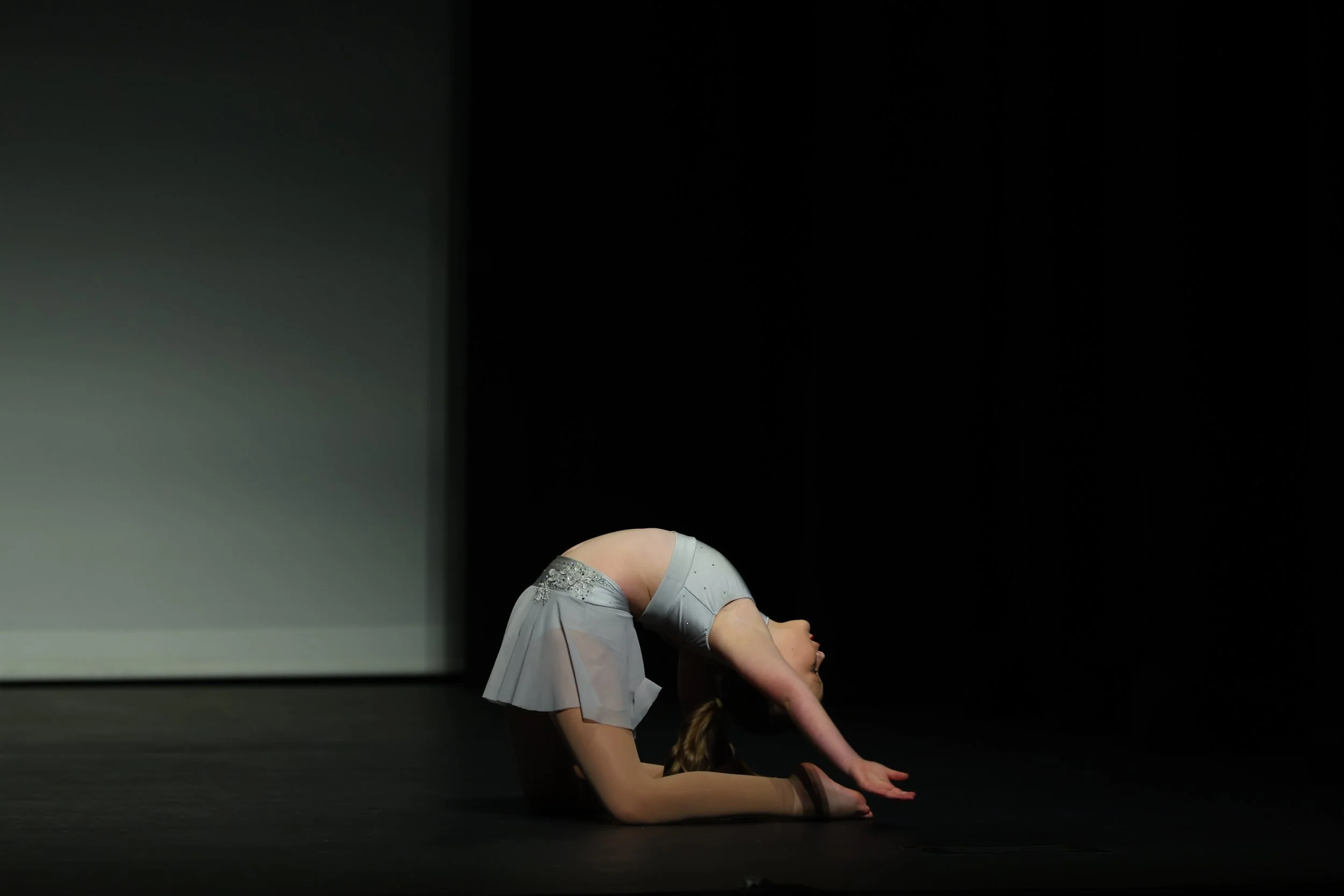 A ballerina in grey costume performing an artistic pose on stage with a black background.