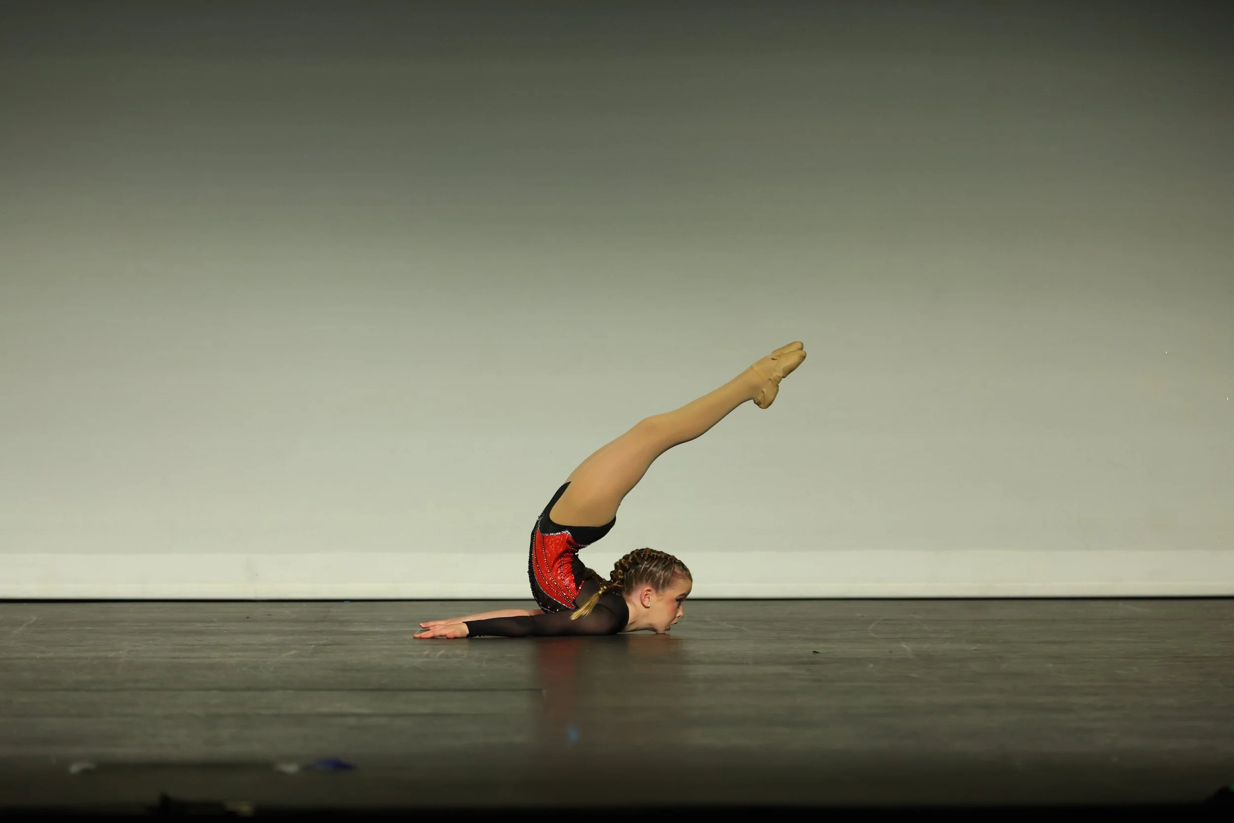 A young girl performing an acrobatic pose on a stage, lying flat with her arms extended and her legs raised vertically, wearing a red and black costume with long hair braided.
