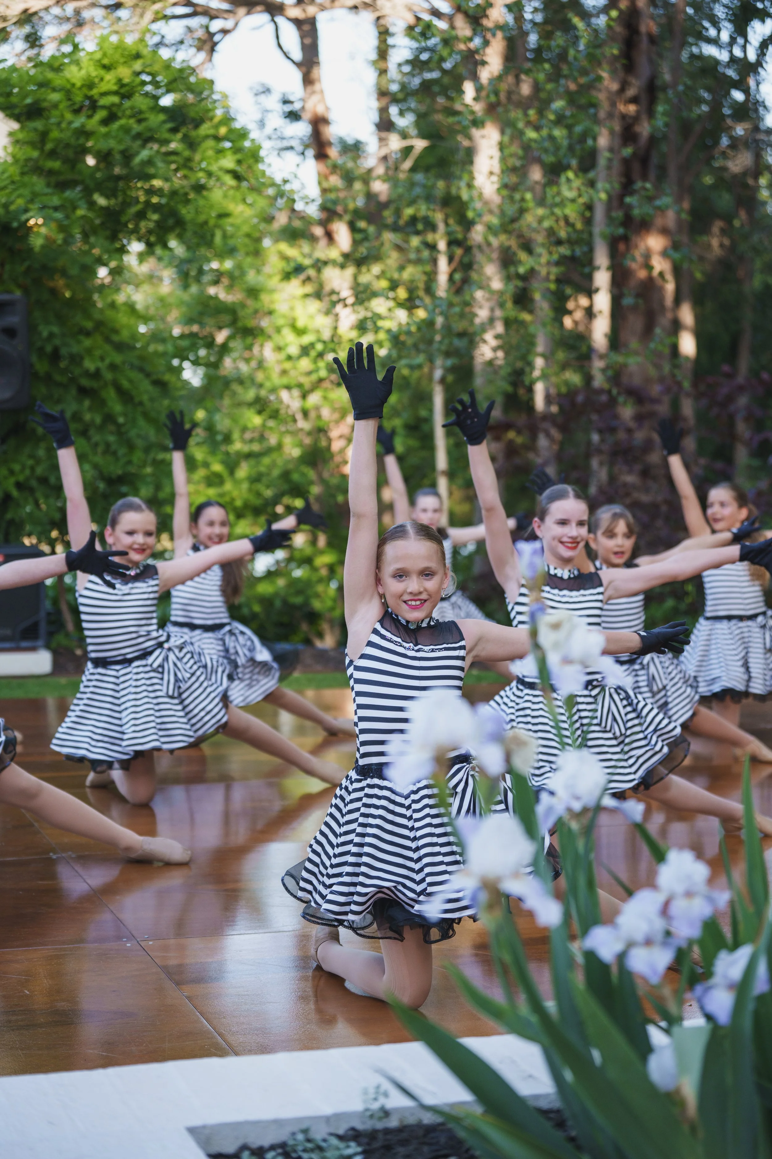 A group of young female dancers dressed in black-and-white striped dresses perform a choreographed routine outdoors on a wooden stage surrounded by trees. Some dancers wear black gloves and black shoes, and they have their arms extended outward with 