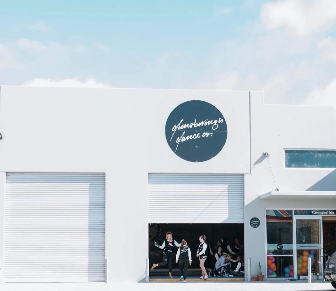 A white building with a black circular sign that reads 'Dunbarough Dance Co.' in white cursive. The building has a large open garage door with dance students inside. To the right, there's a glass door with colorful balls inside visible through the wi