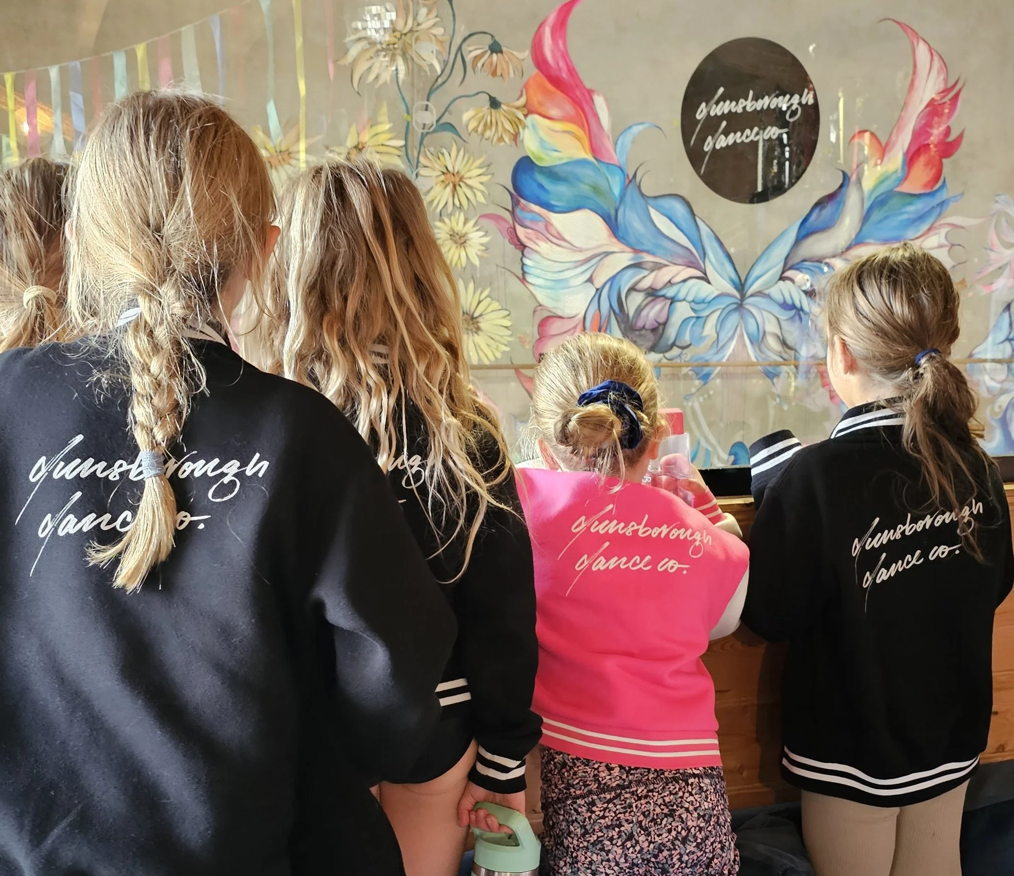 A group of people, mostly children, standing indoors near a colorful mural with flowers and abstract designs. Some are wearing black sweatshirts with white cursive writing. The scene appears to be in a dance studio or classroom.