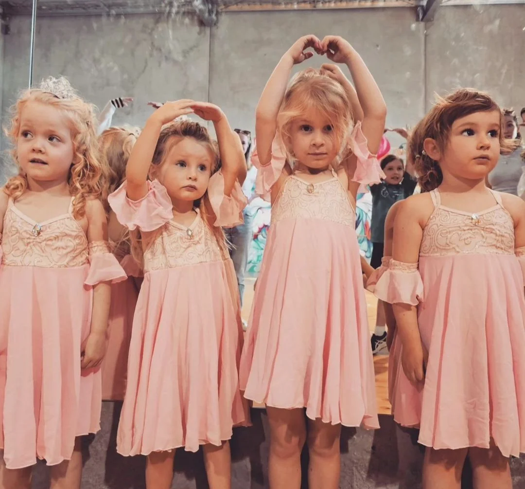 Four young girls dressed in pink dresses standing in a line, some with their hands raised above their heads, at what appears to be a dance or performance event.