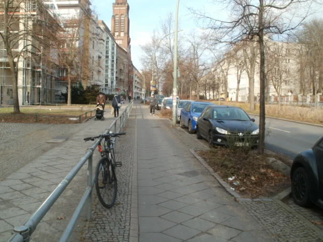 Bike lane in Berlin - separated from foot traffic by a fence and motor traffic by trees and parked cars. Yes, there is a pedestrian walking there...