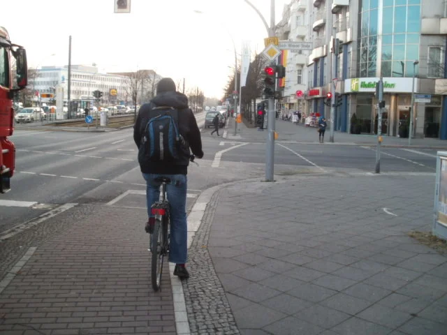 Waiting at a stop light in Berlin. Notice how the bike lane veers a little right to be back on the sidewalk - completely separated from both motor traffic and foot traffic. The small triangle just to the left of the bike lane is a left turn holding lane for bikes. 