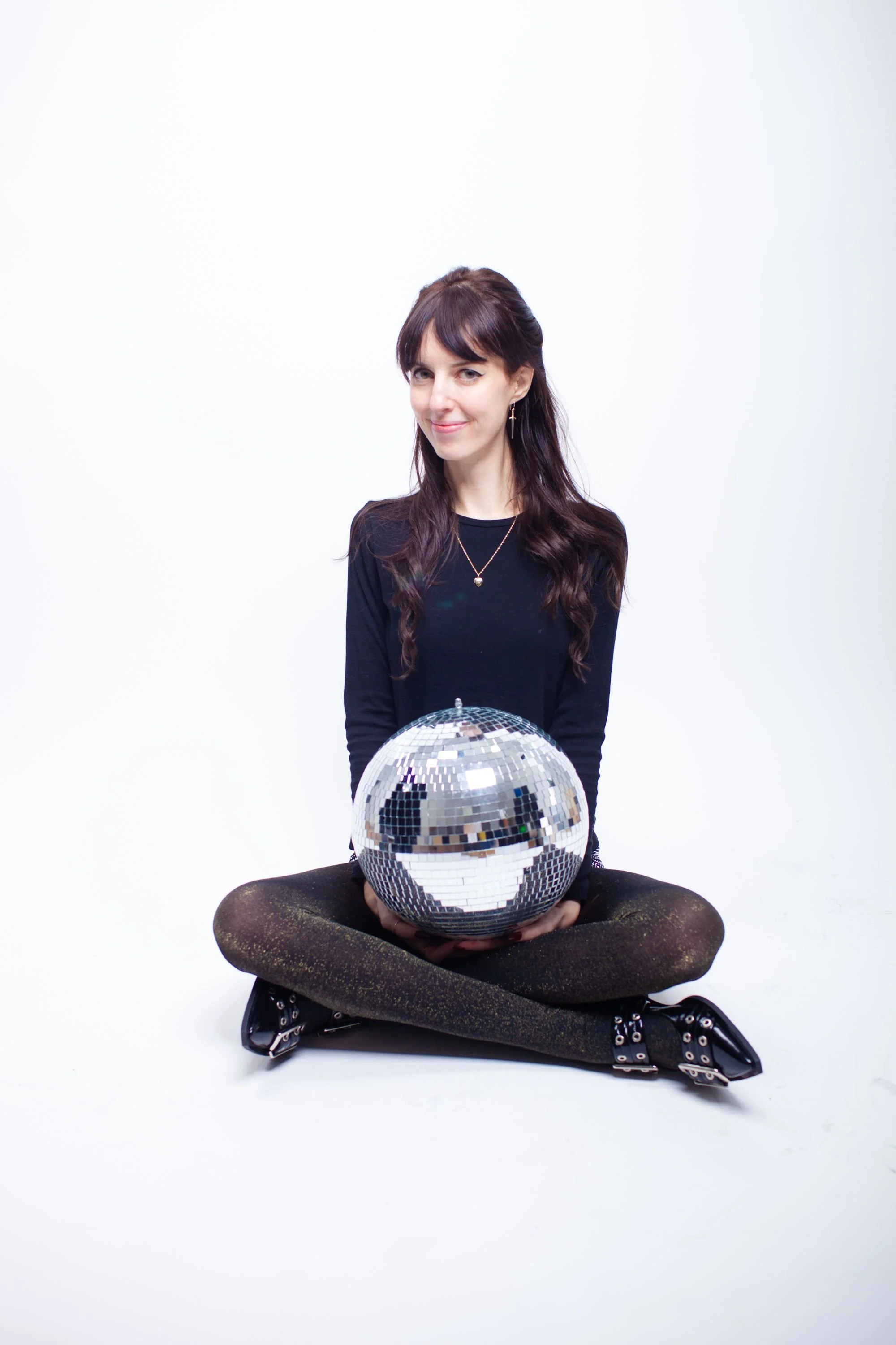 Professional headshot of Madeleine Barnes, a seated woman in a black shirt holding a disco ball smiling confidently against a white background, artistic portraiture.
