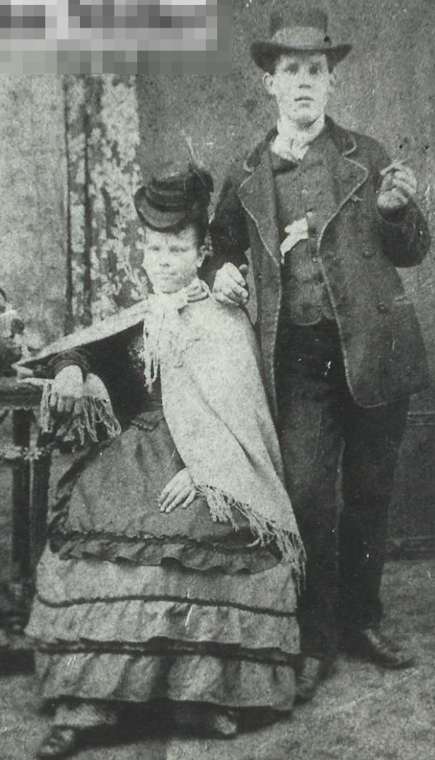 An image of a woman, seated, in ruffled skirts and shawl and a man standing behind in a waistcoat and top hat, flaunting a cigar. Possibly 1870s?