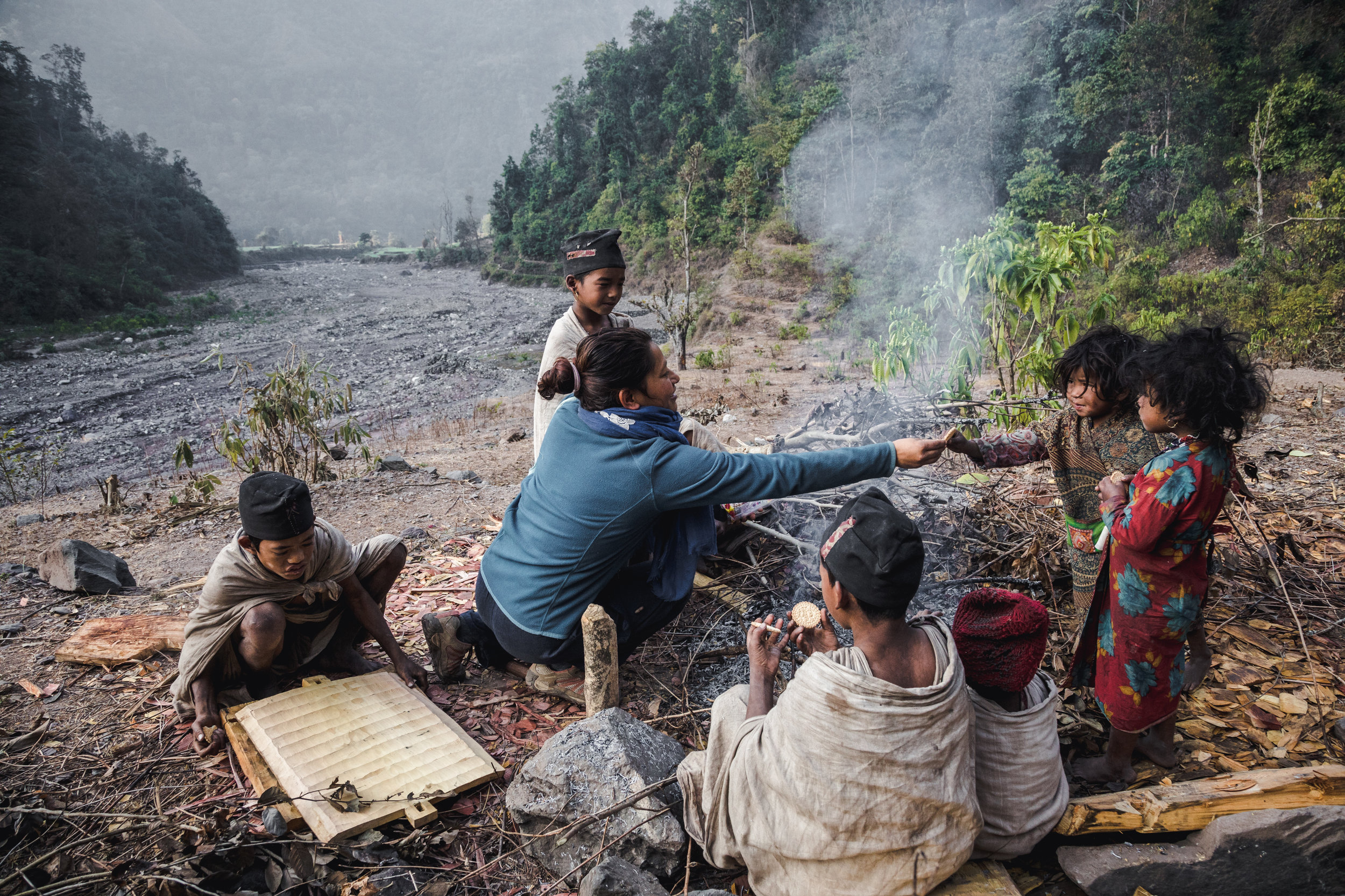 Samjhana handing over fruits and sweets to children sitting on the riverbed next to the jungle.