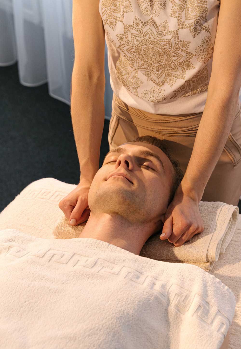  Young man lies on his back with towel covering up to his neck. Female masseuse is preparing to start the masssage. 