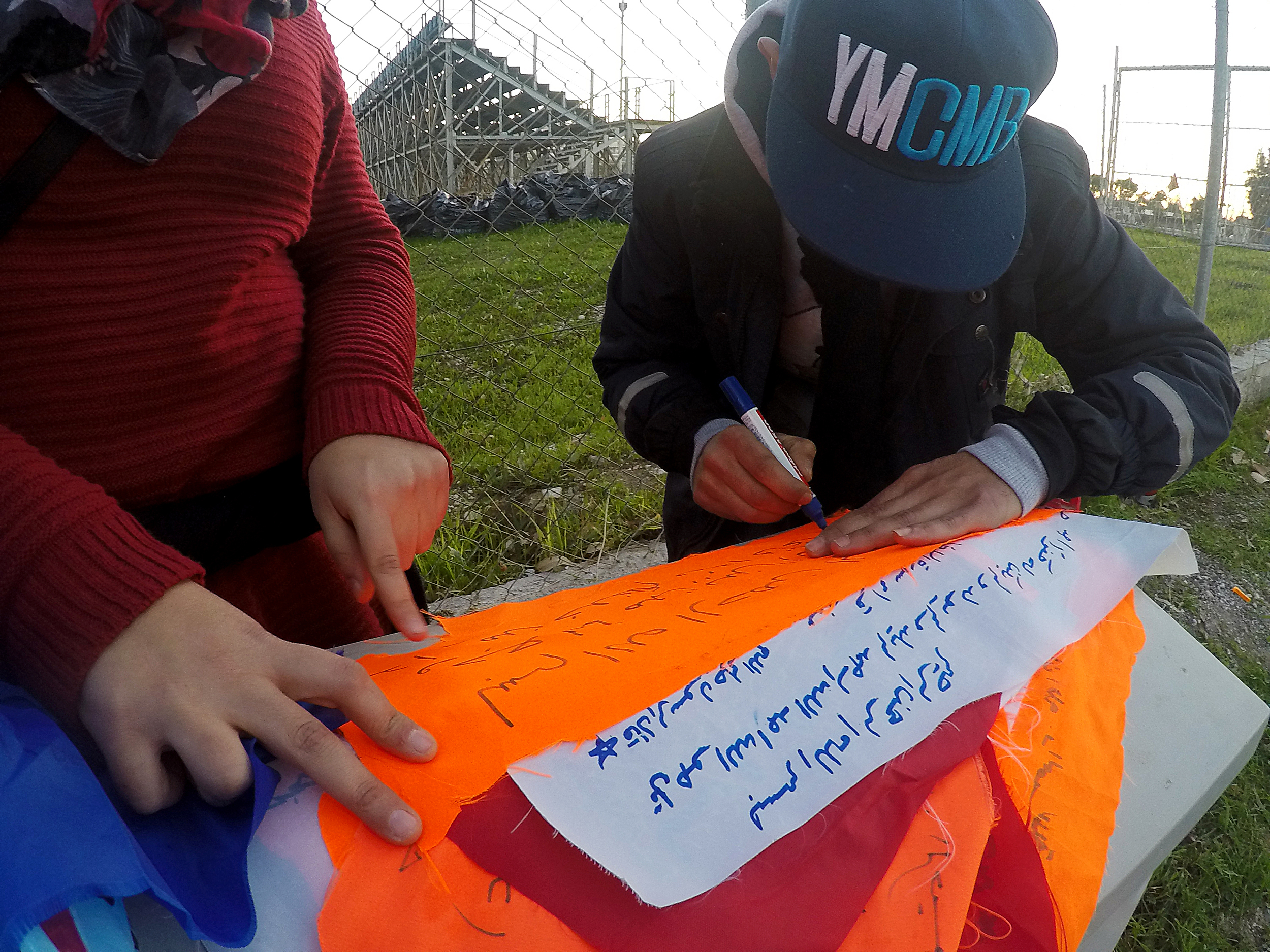 Refugee men writing prayers and protests on life jacket swatches outside of Refugee Camp Tae Kwon Doe, in Athens, 2015. Photo courtesy of artist.
