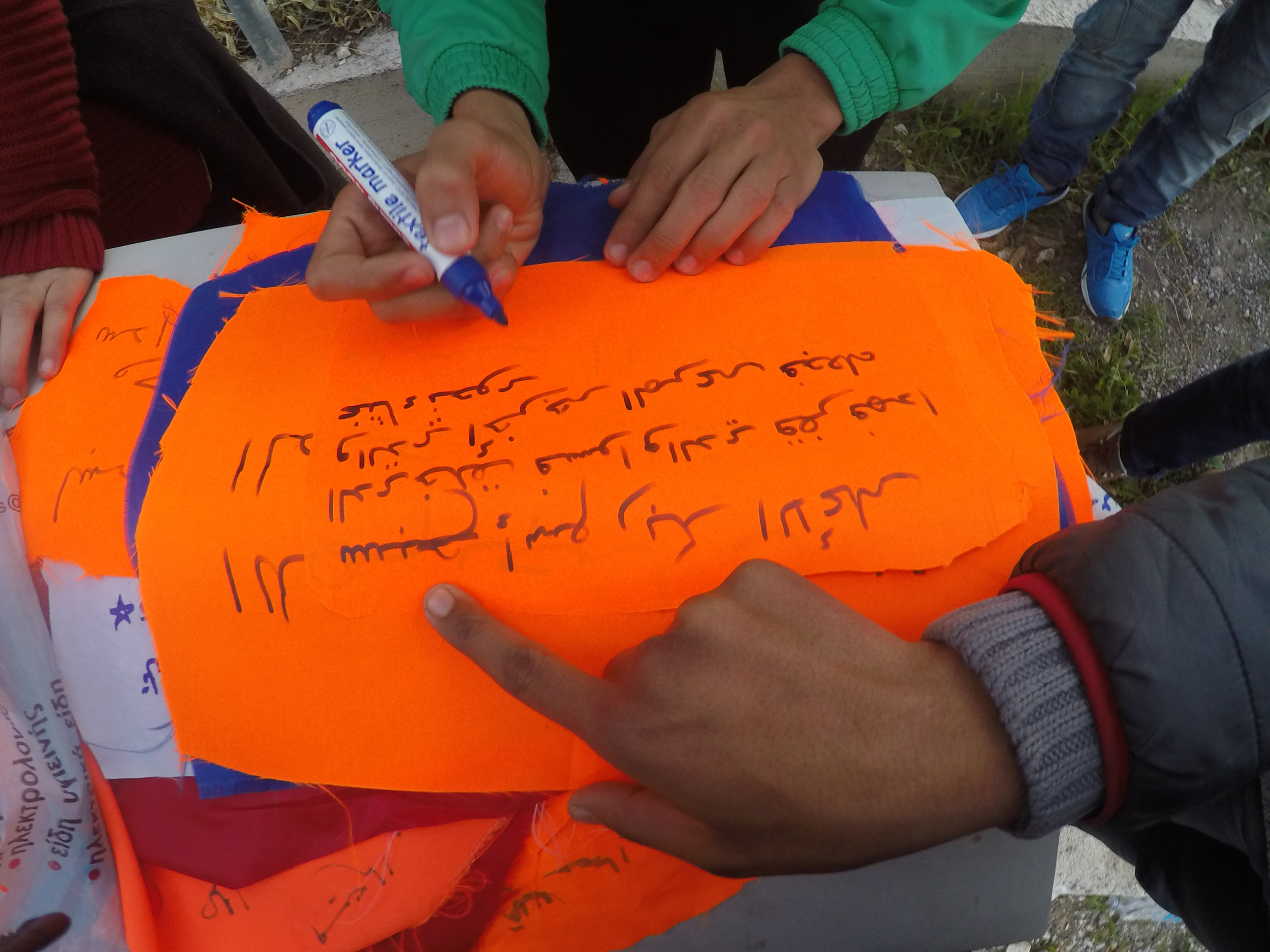 Refugee men writing prayers and protests on life jacket swatches outside of Refugee Camp Tae Kwon Doe, in Athens, 2015. Photo courtesy of artist.