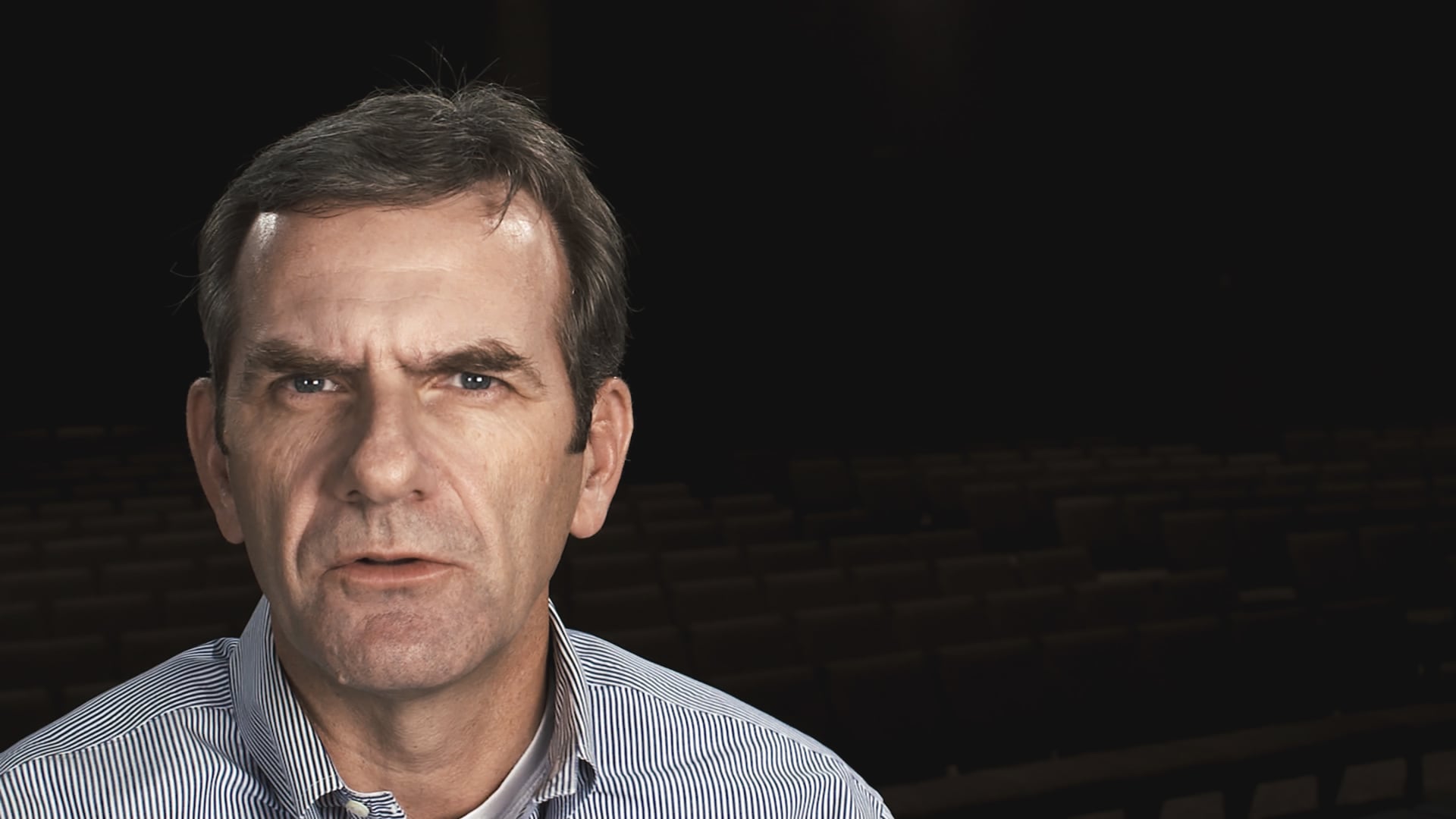You can faintly see the rows of chairs behind him as he speaks directly into the camera.