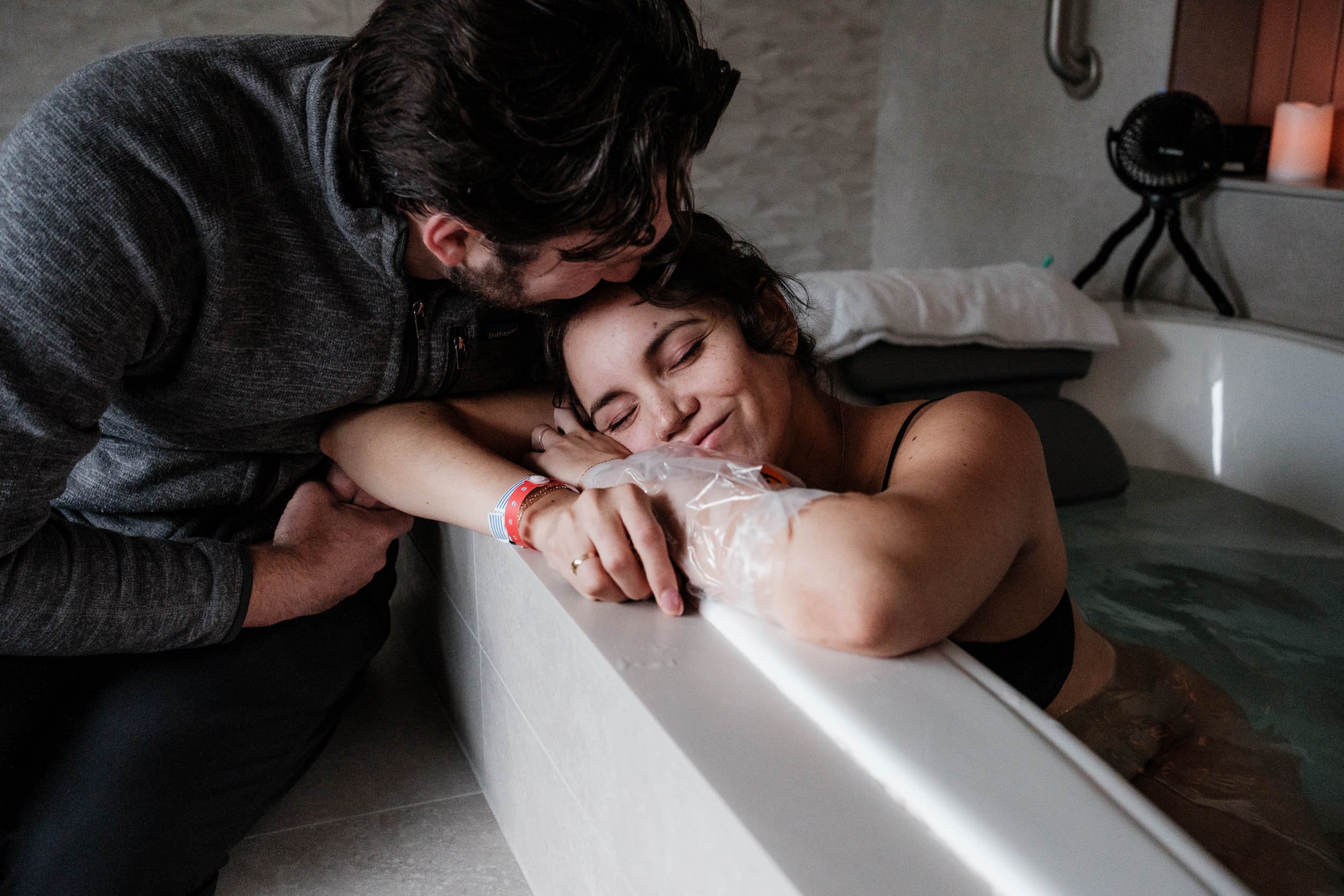 A husband kisses his wife's forehead while she smiles between contractions and sits in a birthing tub.