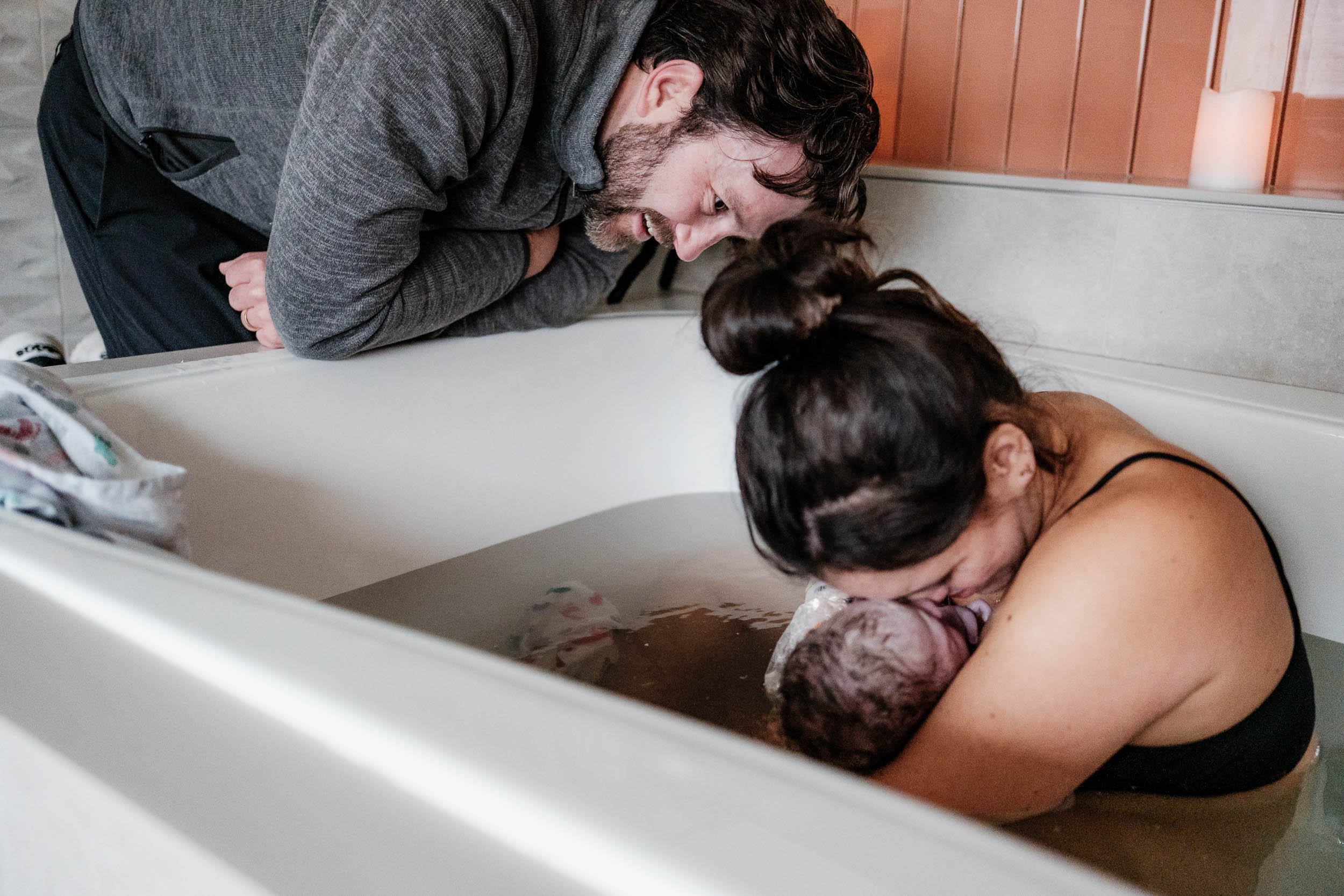 A father leans over the birth tub to take a close look at the baby his wife just birthed and is holding in her arms.