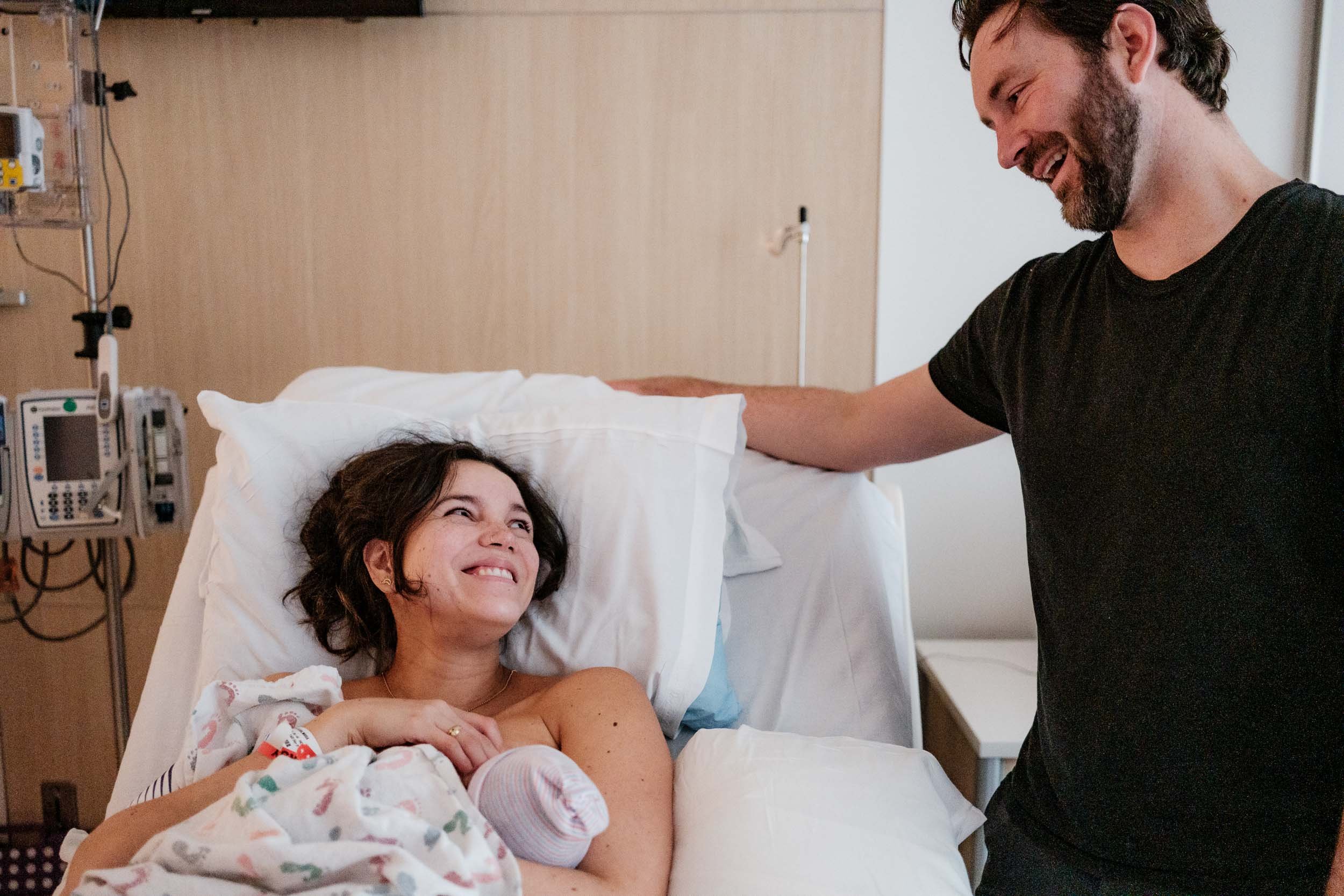 A mother smiles at her husband from the hospital bed with their newborn son in her arms.