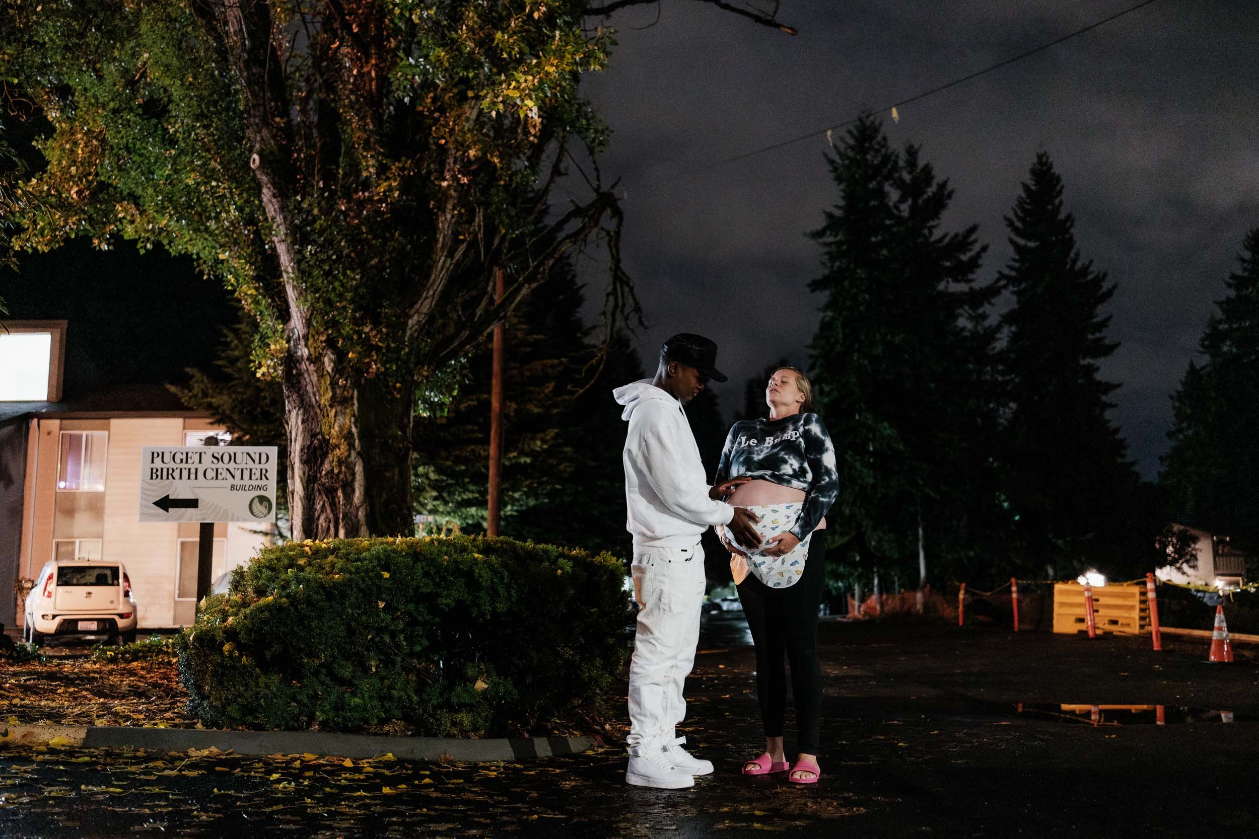 A mother in labor, holds her belly up as she works through a contraction outside under the night sky as her husband touches her belly and supports her.