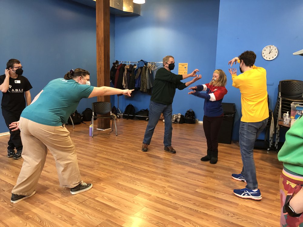 [ID: Teaching Artist Kaylie stands in the center of a circle and animatedly points to Creative Associate, Rachel. Rachel, flanked by Dennis and Joe work together to make an alligator.] 