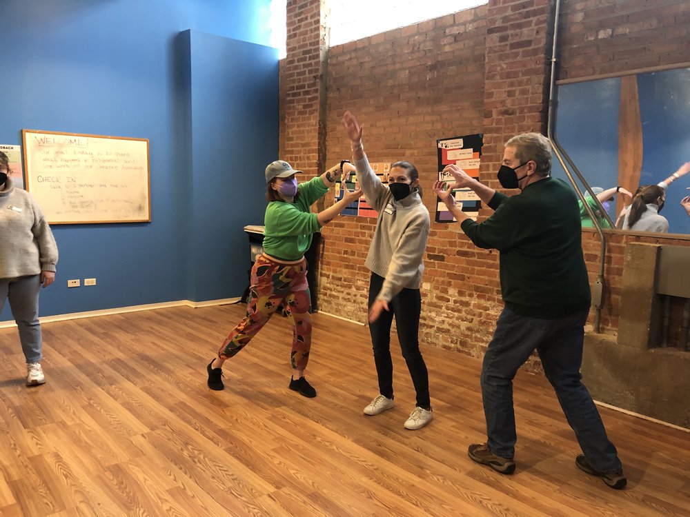  [ID: a rehearsal studio with a bright blue wall and exposed brick. Development Manager Michelle reaches her arms out in front of her to make the snapping jaws of an alligator. On either side of her, Board members Mary Kate and Dennis make small circ