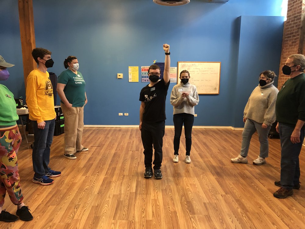  [ID: the group makes 2 lines facing each other. In the center Creative Associate Sam raises his arm proudly in the air. Michelle stands just behind him to drop in a line.] 
