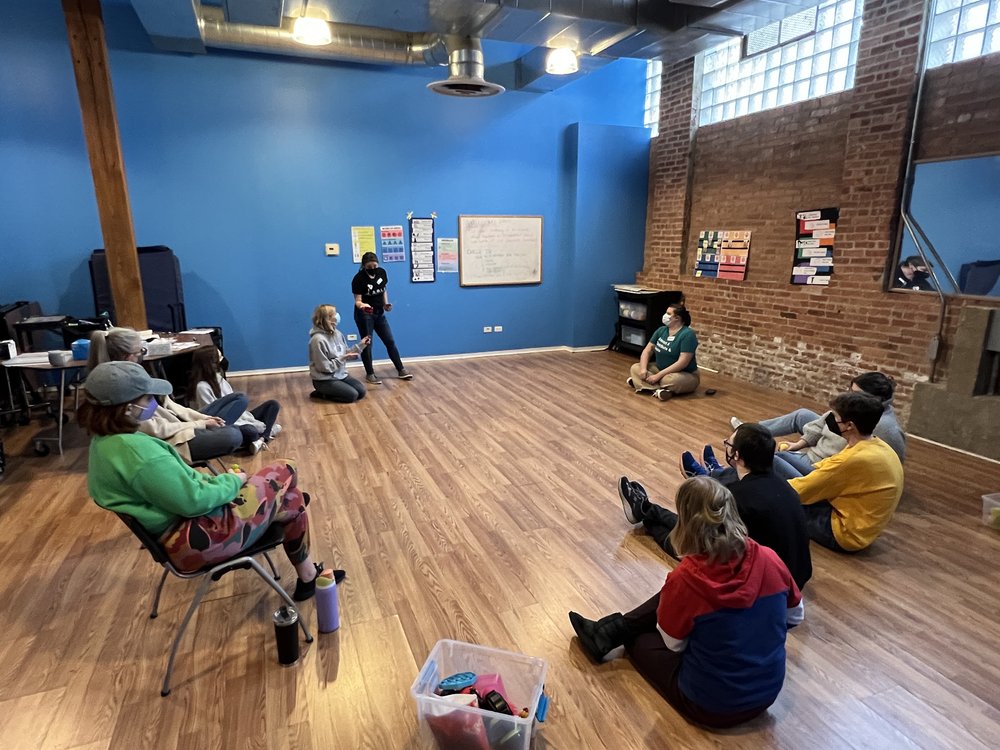  [ID: the group of 11 people circle together for a check out to reflect on the session. Some are seated on the floor and others in chairs. Executive Artistic Director, Katie stands at the far end. She hands a rose to Teaching Artist, Arin, to start t