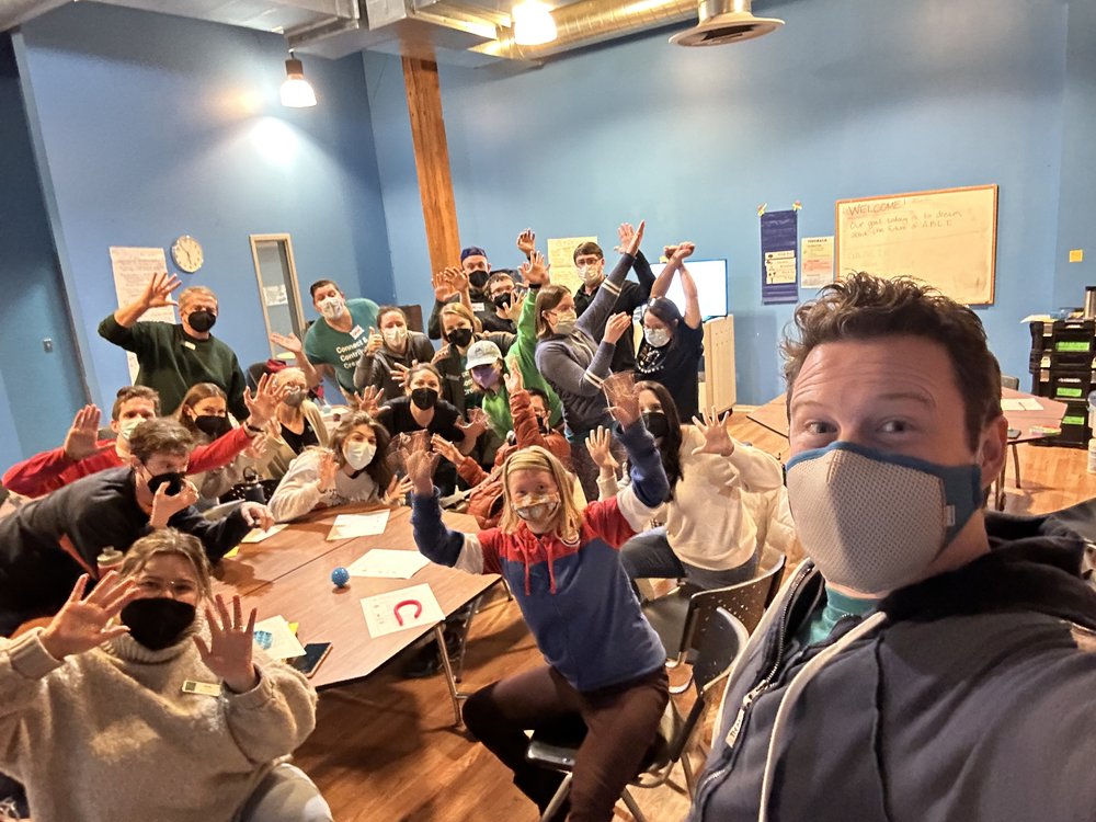  [ID: Teaching Artist Lawrence snaps a selfie of all 21 attendees at our Strategic Planning Session. The group clusters together around a large wooden table in a room that is painted bright blue. Some people sit and others stand. All are wearing face