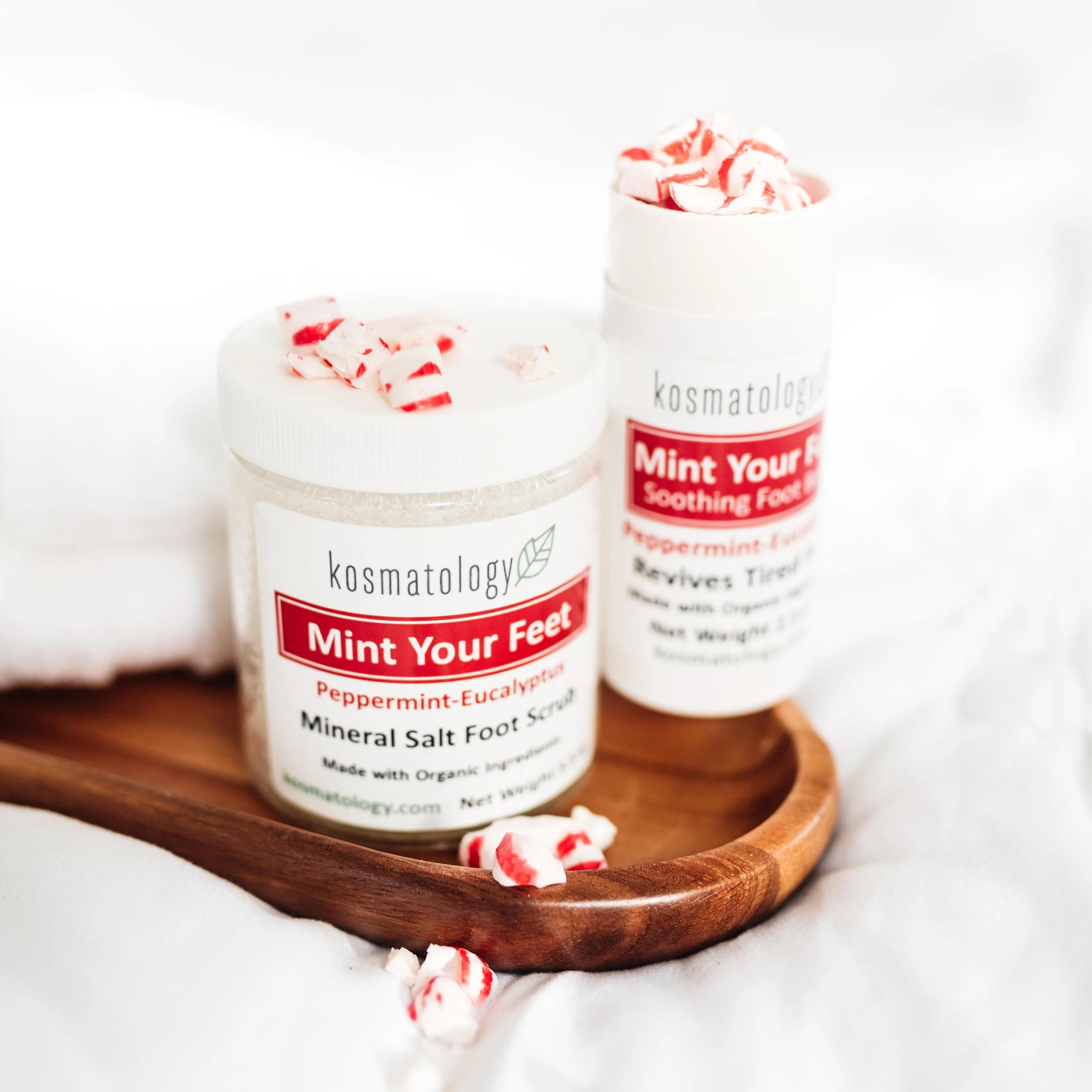 Kosmatology Mint Your Feet foot balm and scrub on a wooden tray with peppermints scattered.