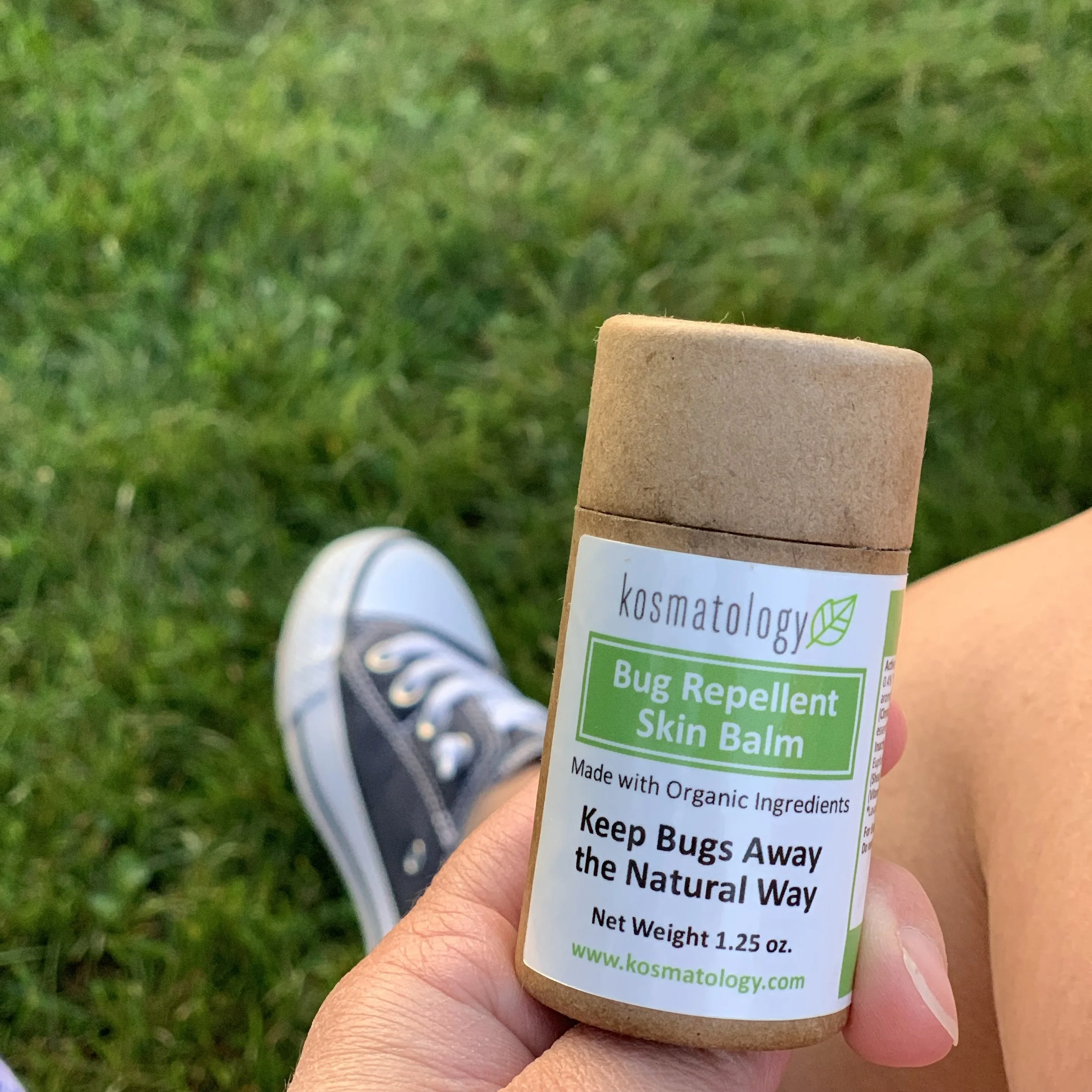 Kosmatology insect repellent balm being held with grass and sneakers in the background