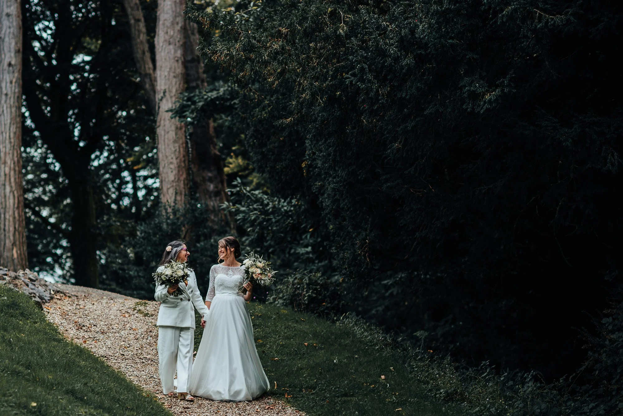 Two brides walking on a forest path, one wearing a white dress and the other a white suit, holding bouquets and smiling at each other.