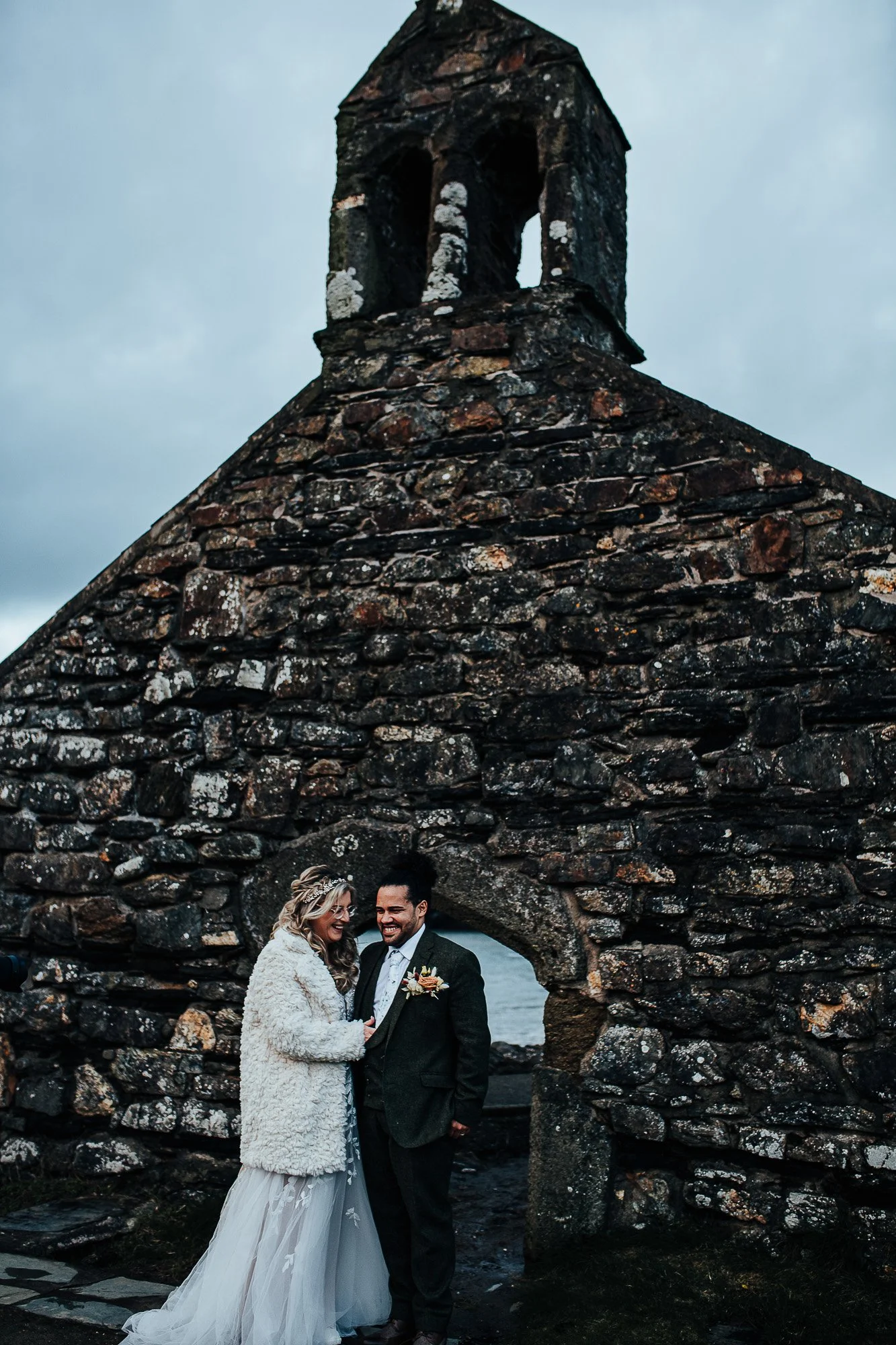 A bride and groom standing in front of an old stone chapel with a small arched window. The bride is wearing a white wedding dress and a fur coat, while the groom is in a dark suit. They appear to be smiling and looking at each other. The sky is overc