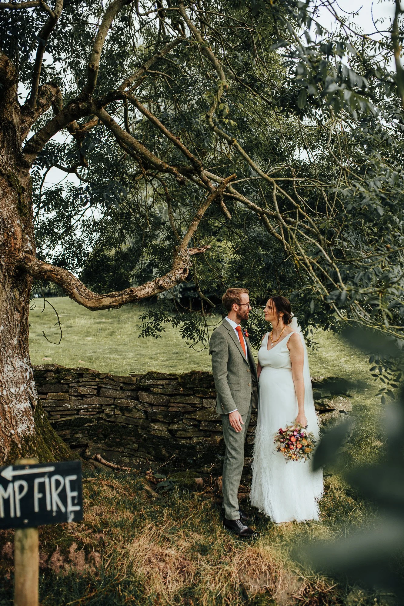 Bride and groom standing under a tree with a stone wall in the background; bride holding a bouquet and they are looking at each other.