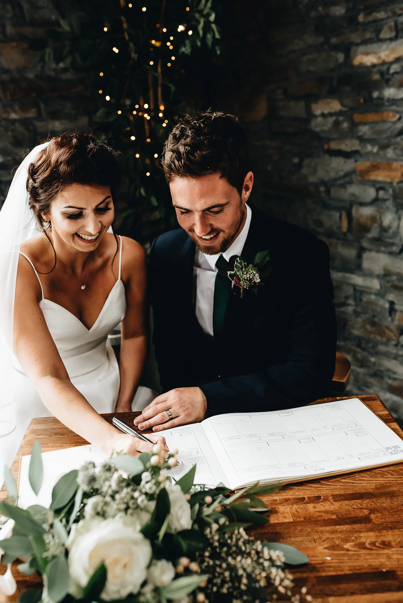 Bride and groom signing wedding register at a wooden table with flowers, smiling.