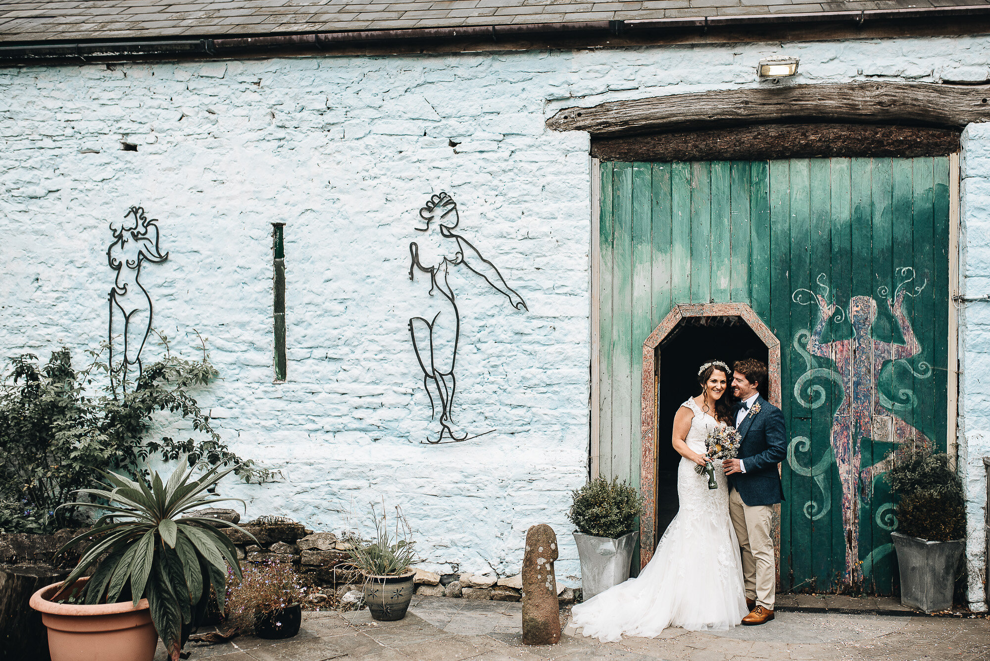 Bride and groom smiling in front of a rustic, painted barn door with artistic wall designs. The bride is wearing a white wedding dress, and the groom is in a suit. Potted plants and abstract figures are painted on the surrounding walls.