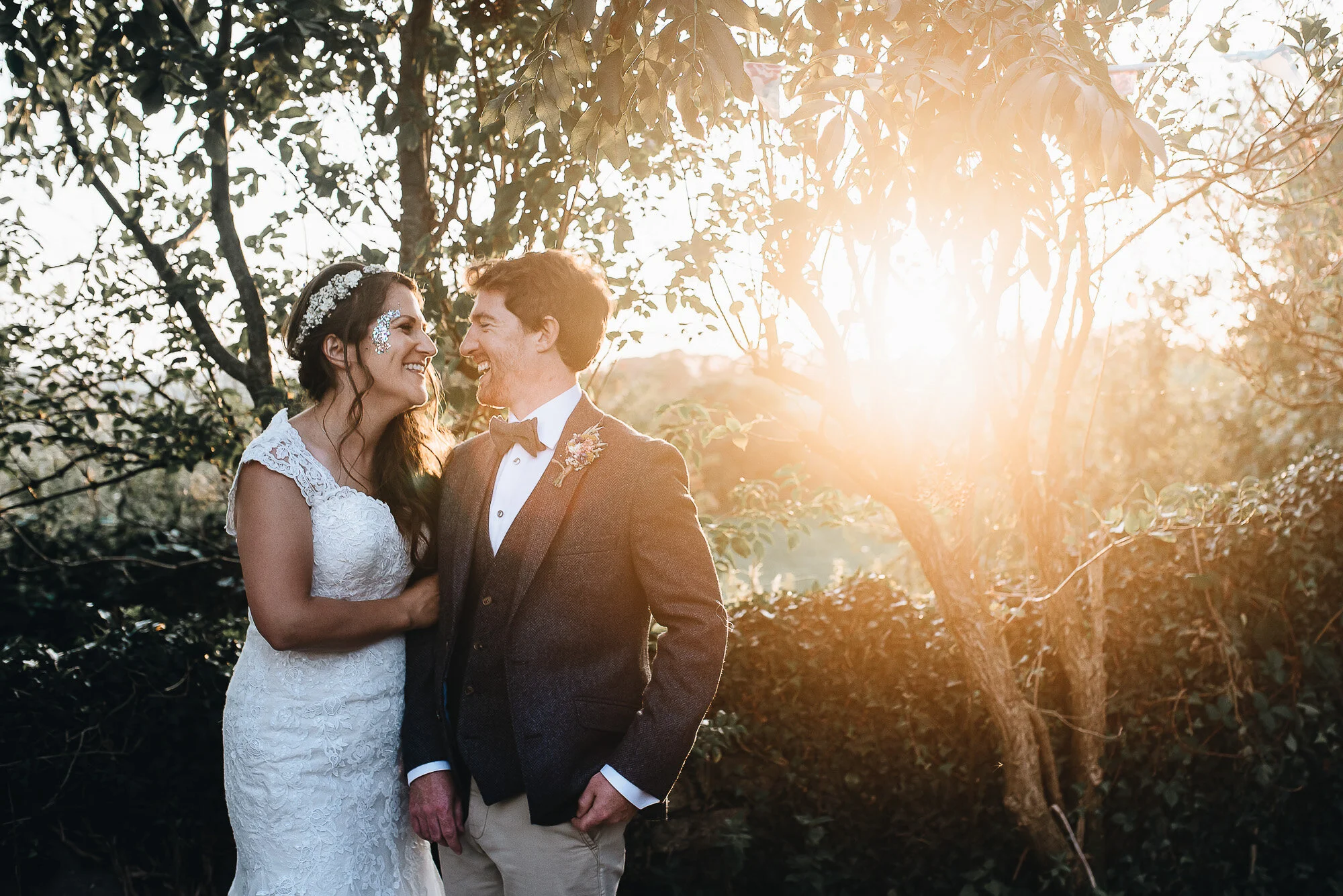 Bride and groom smiling at each other outdoors with sunset light through trees.