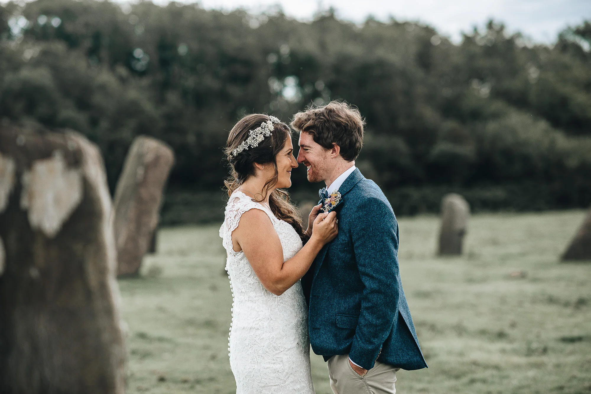 A bride and groom embracing outdoors with standing stones in the background.