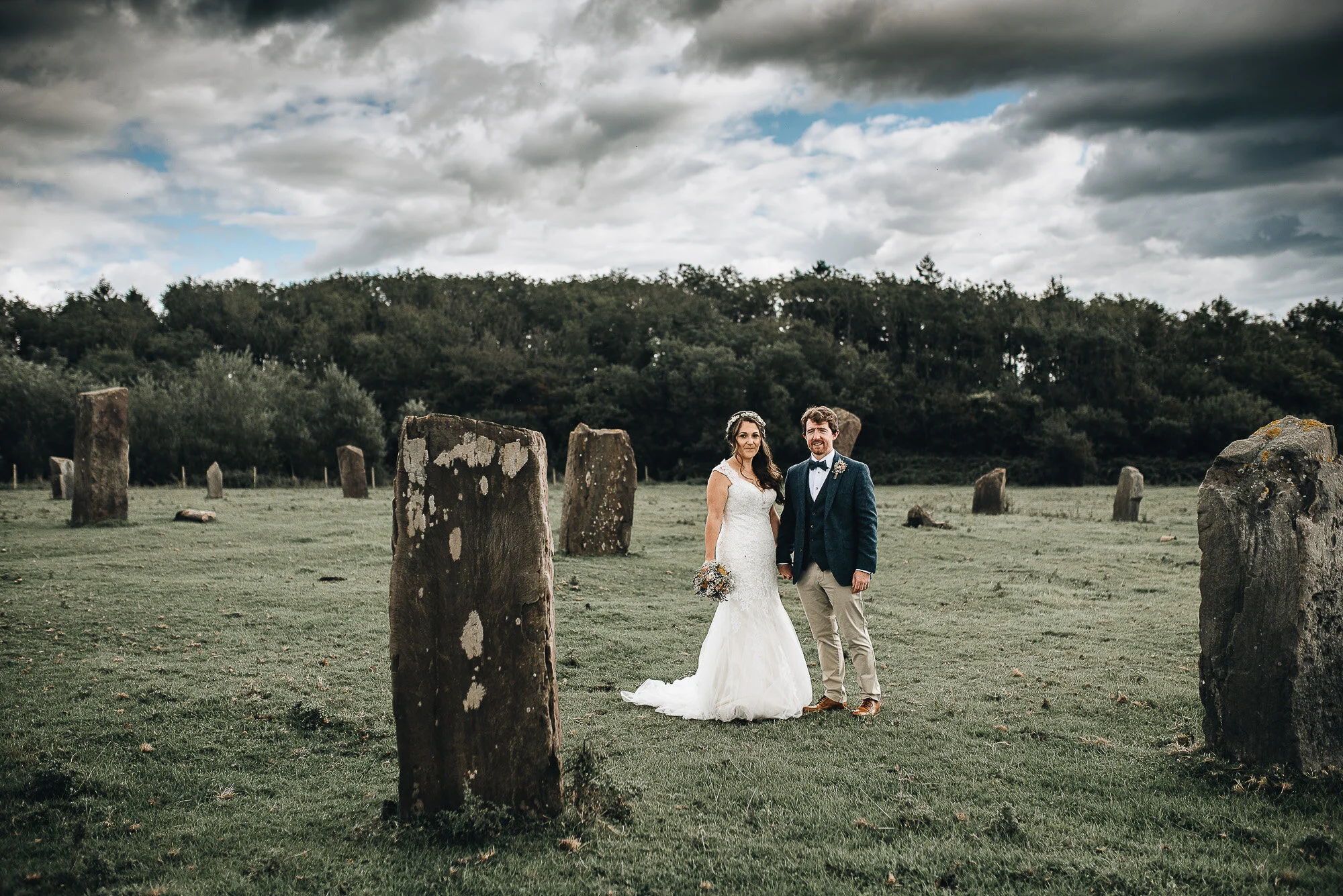 Bride and groom standing among ancient stone monoliths, overcast sky, grassy field, dense tree line in background.