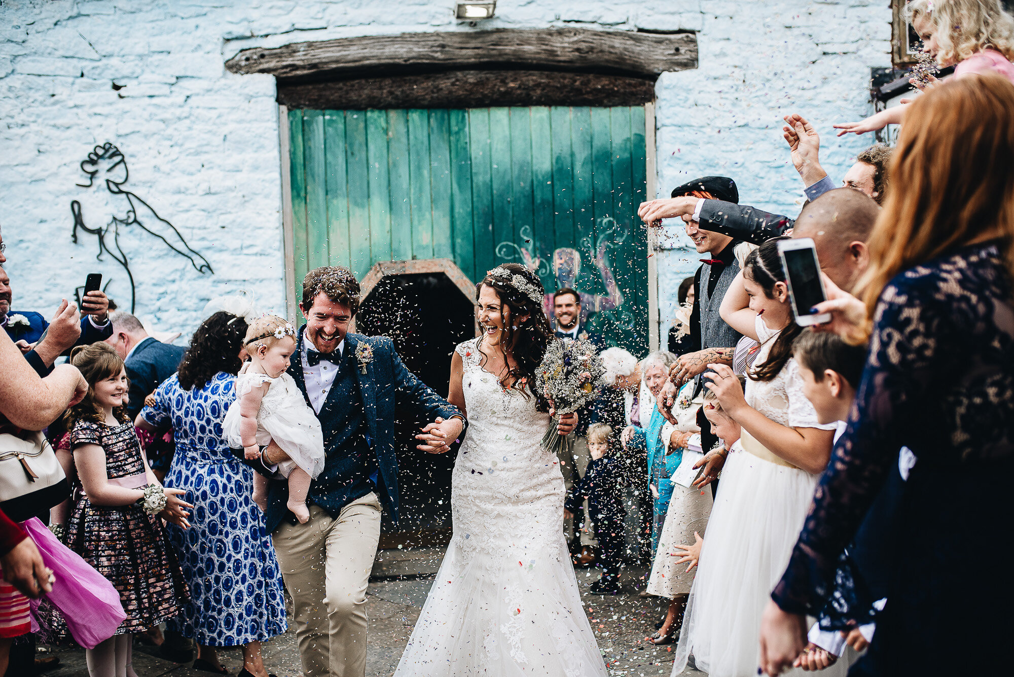 Bride and groom walking through guests throwing confetti outside a rustic building, holding a baby.