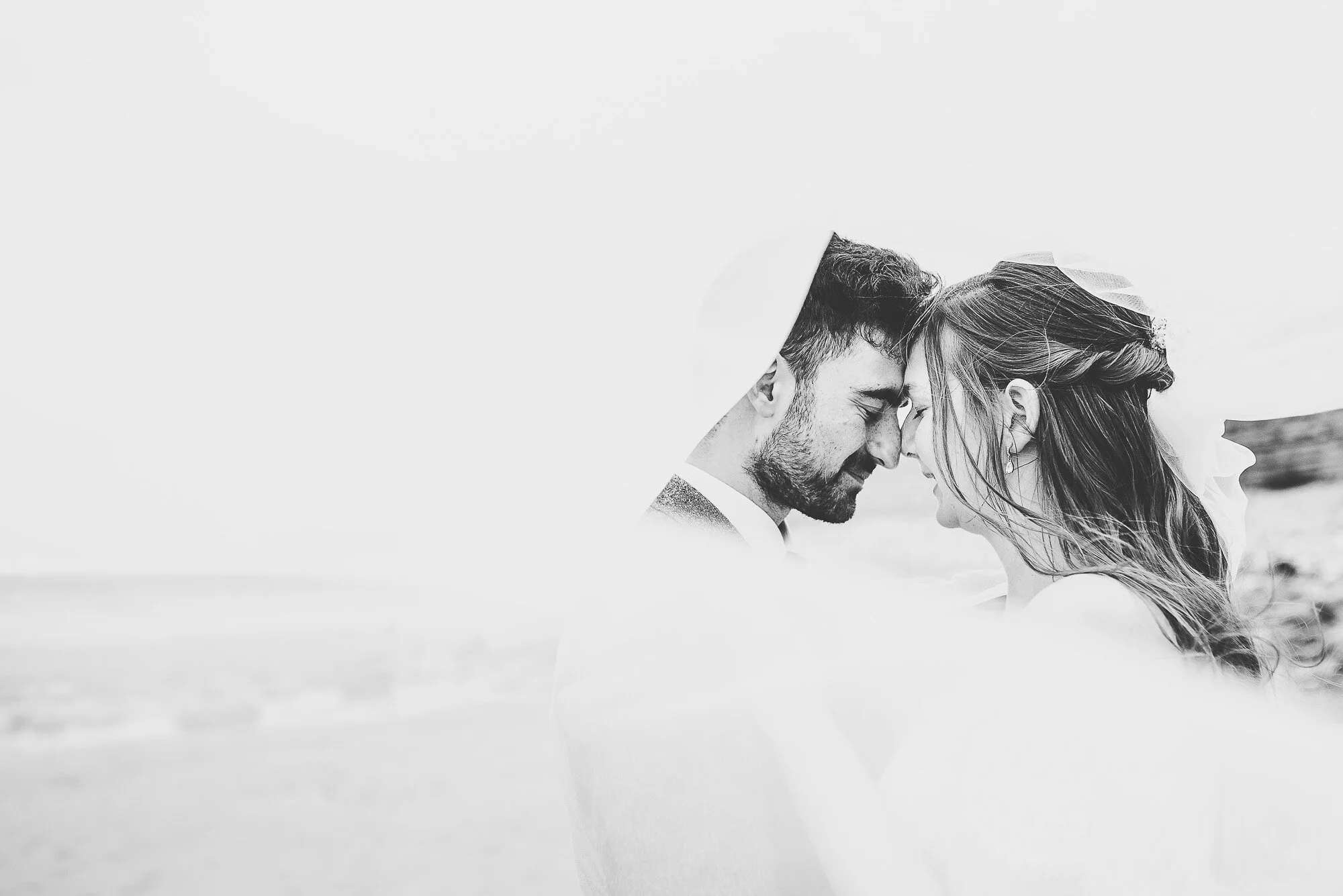 Bride and groom in close embrace under a veil, black and white photo.